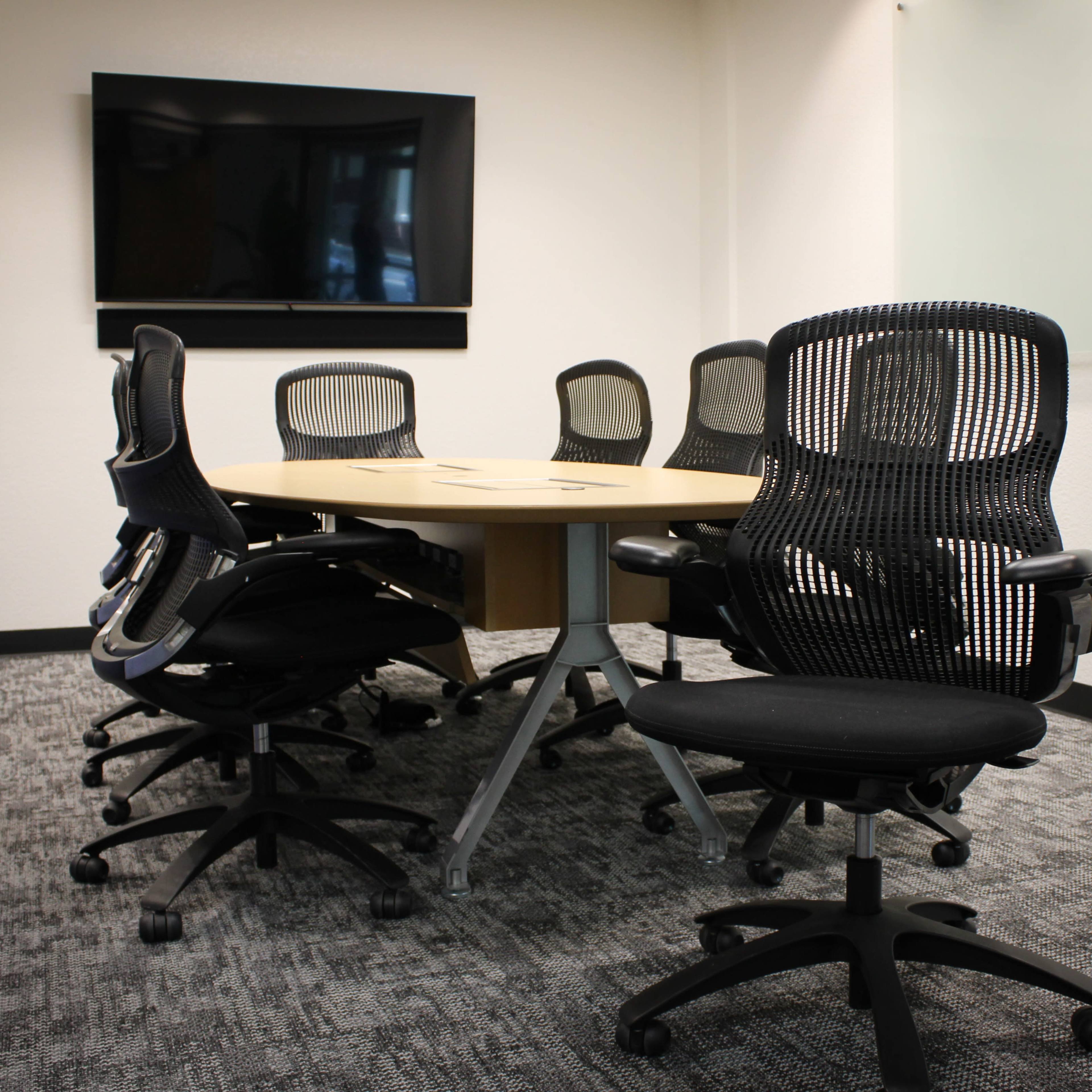 A circular conference table surrounded by black ergonomic chairs is situated in a well-lit meeting room with a television screen mounted on the wall.