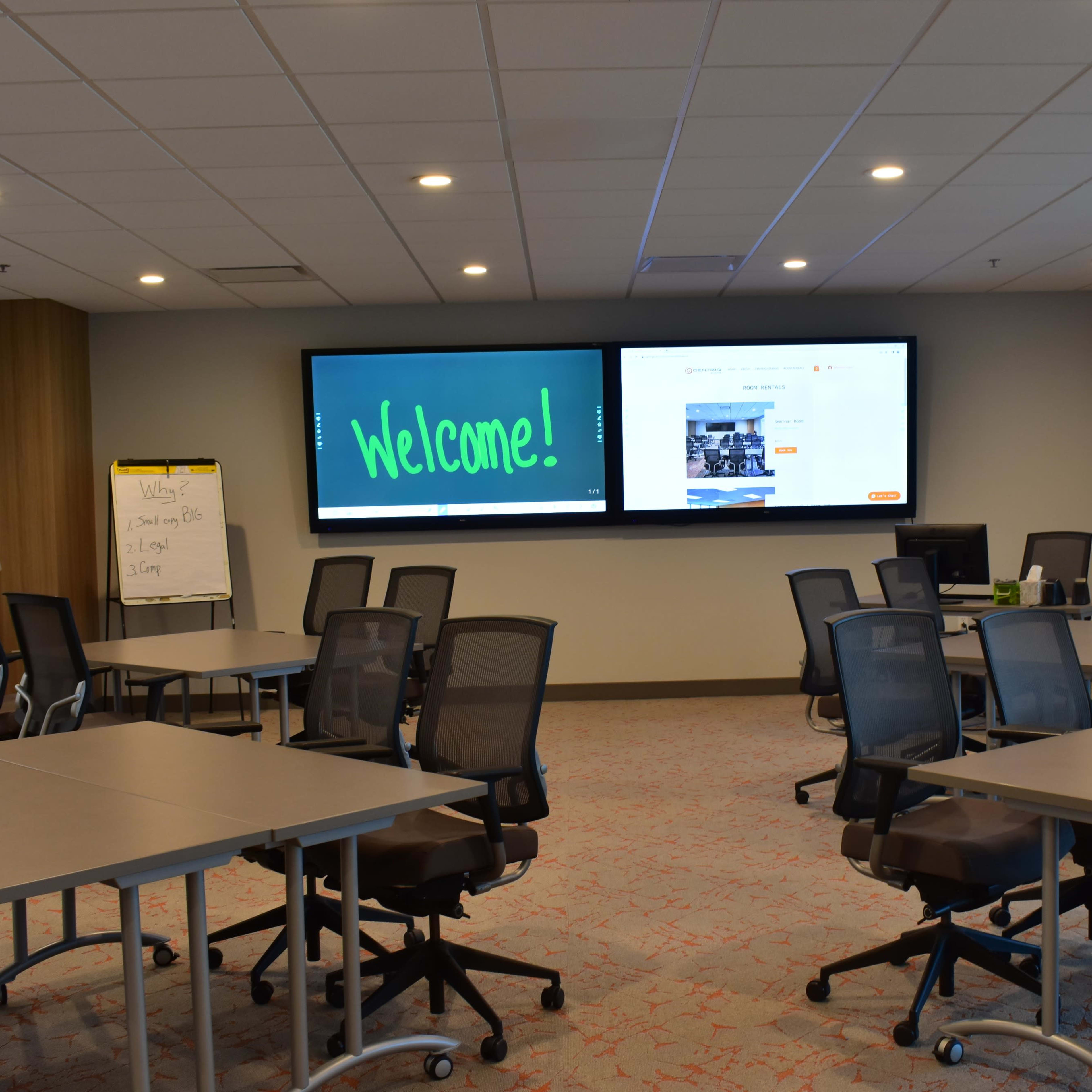 A modern conference room with tables arranged for a meeting, featuring two large screens displaying a welcome message and additional content.