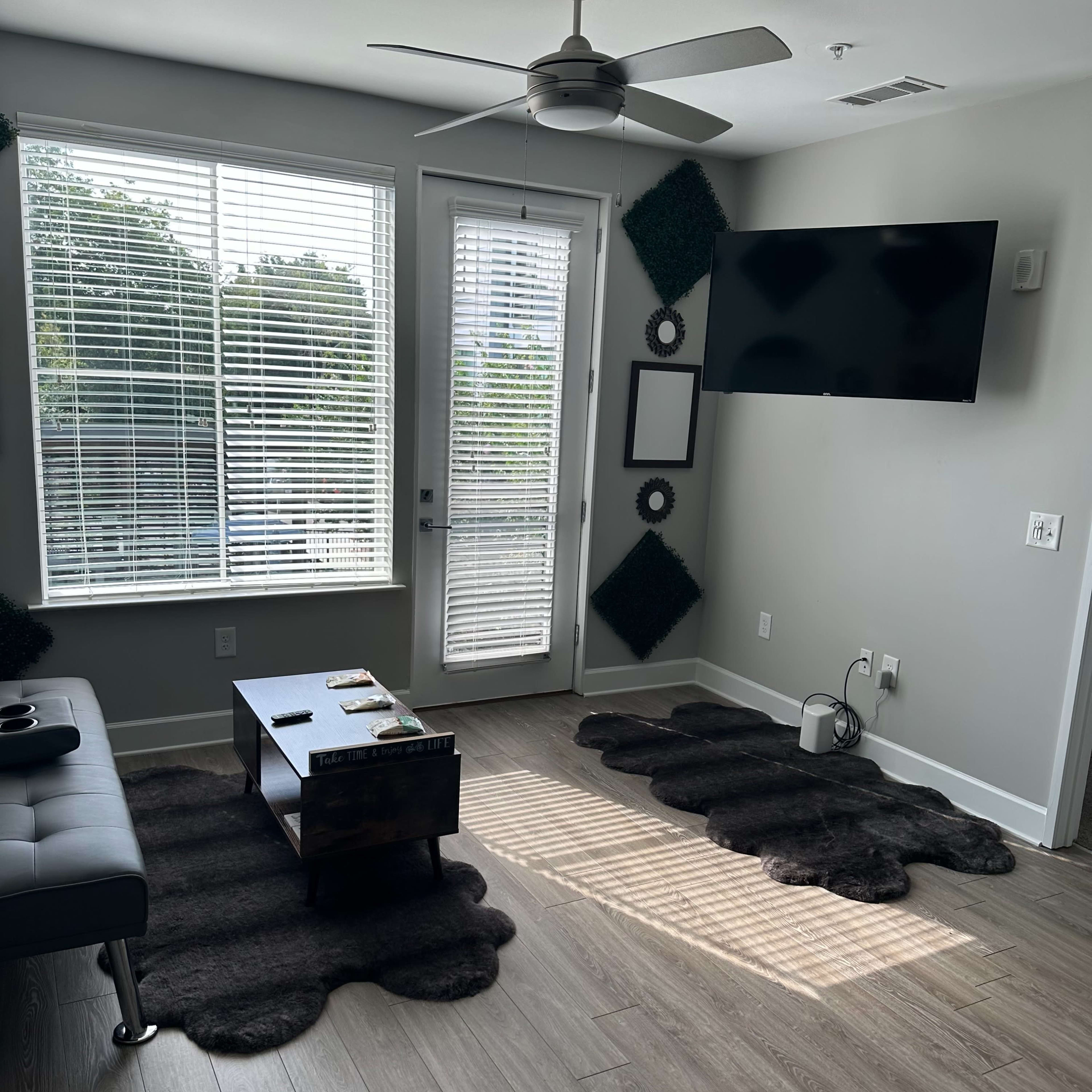 A modern living room features a dark rug, a coffee table, a wall-mounted TV, and large windows with blinds.