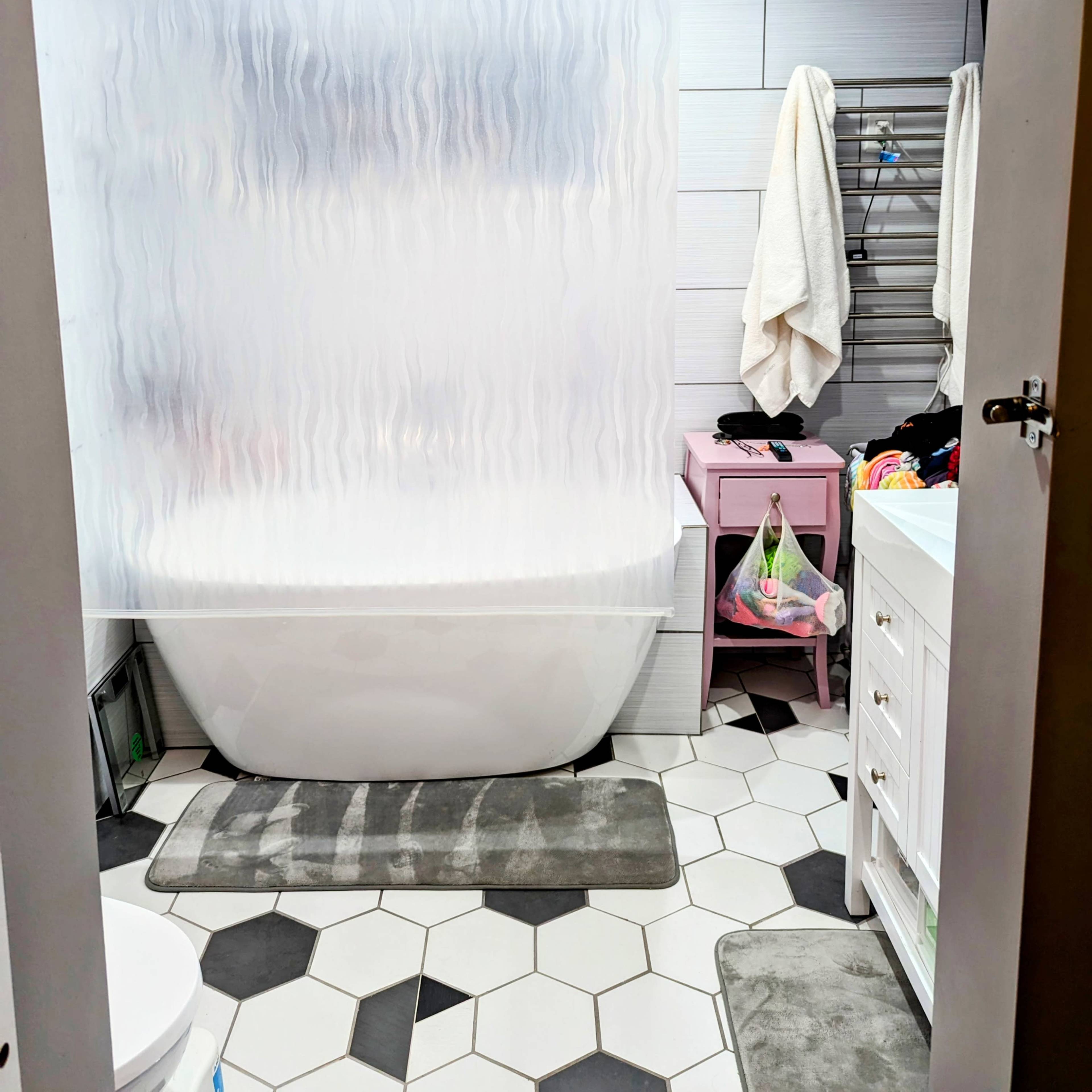The bathroom features a freestanding bathtub separated by a wavy shower curtain, with a hexagonal tiled floor and various bathroom accessories.