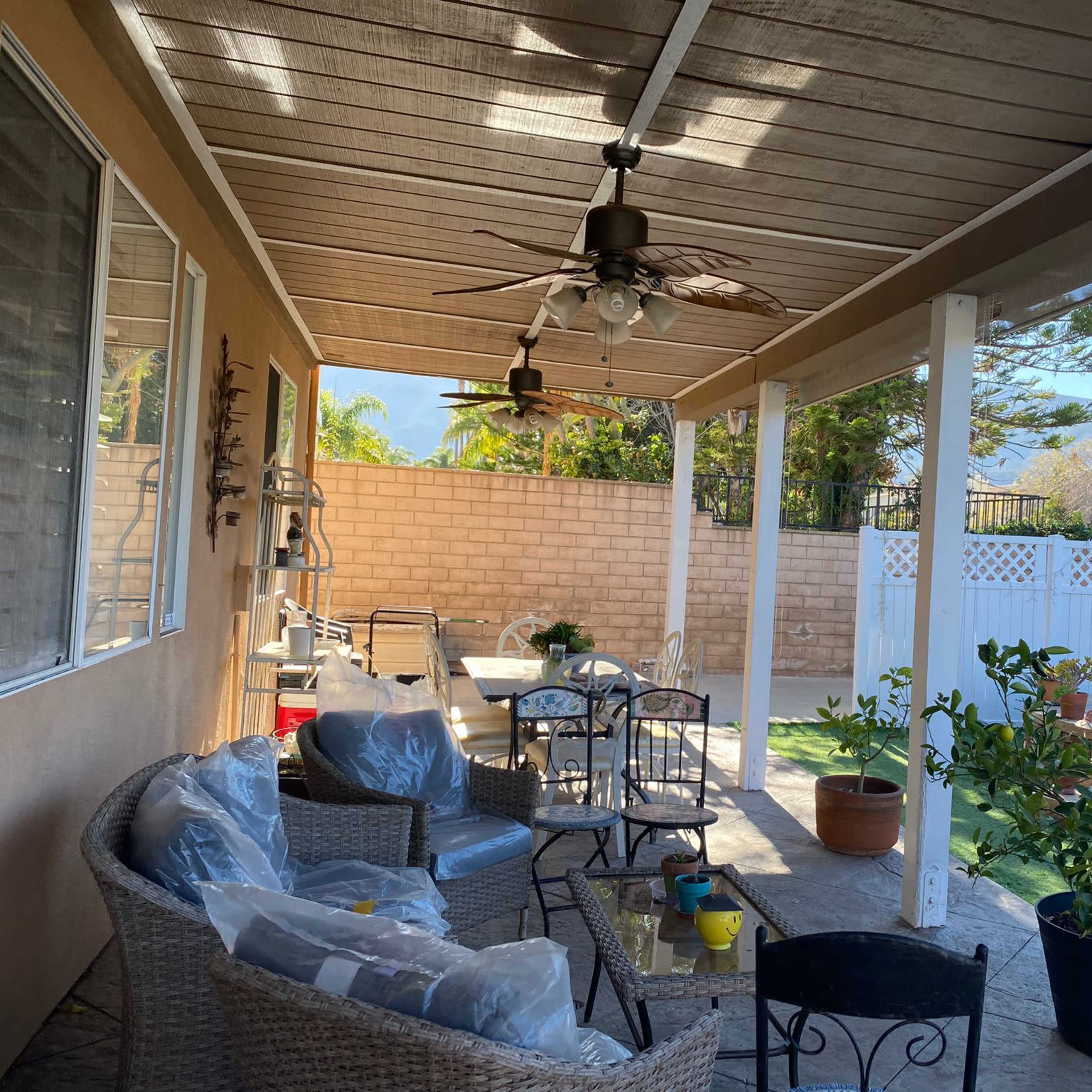 The photo shows a covered patio area with wicker furniture, a ceiling fan, and a view of a landscaped yard featuring a stone wall and a mountain backdrop.