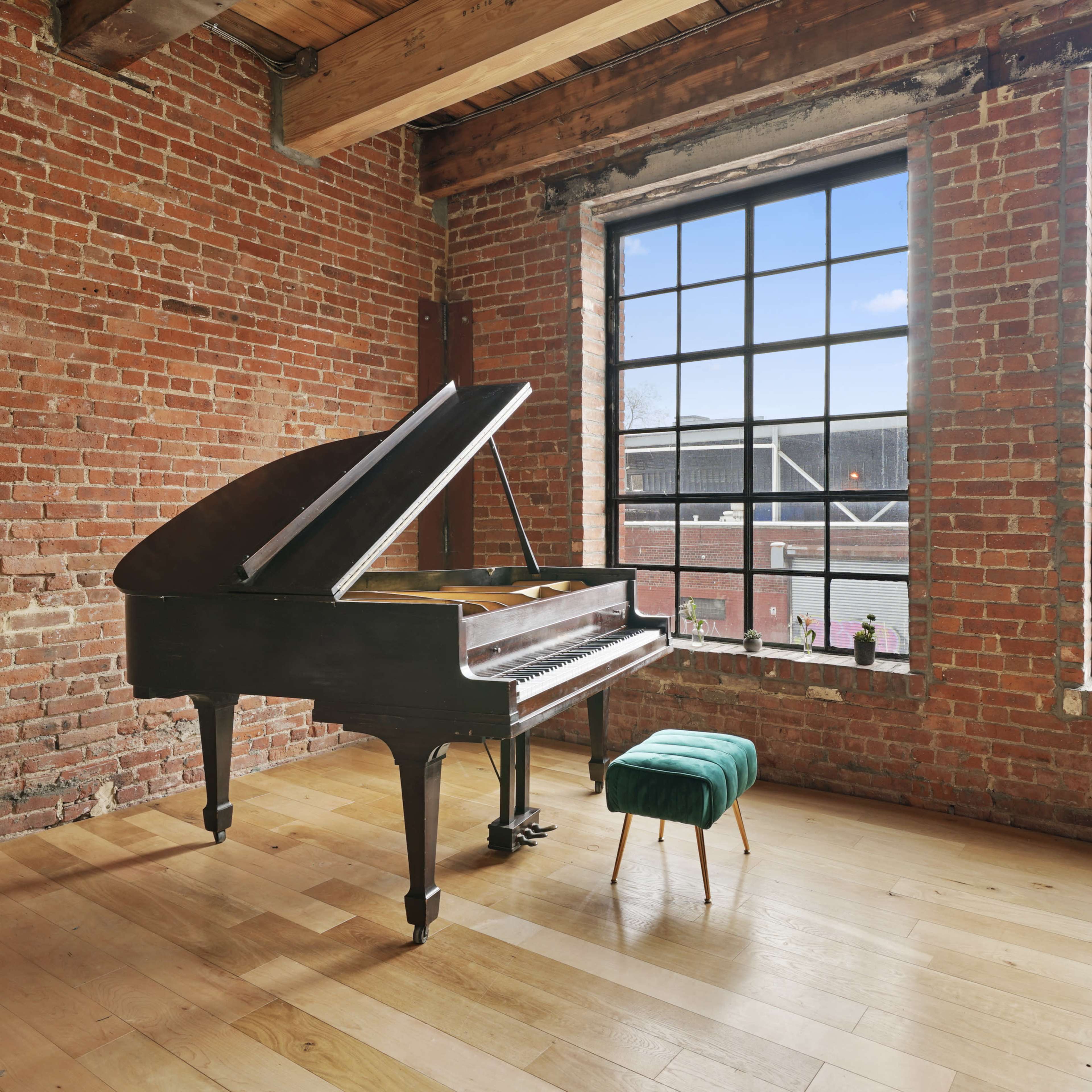 A grand piano sits next to large windows in a room characterized by brick walls and wooden flooring.