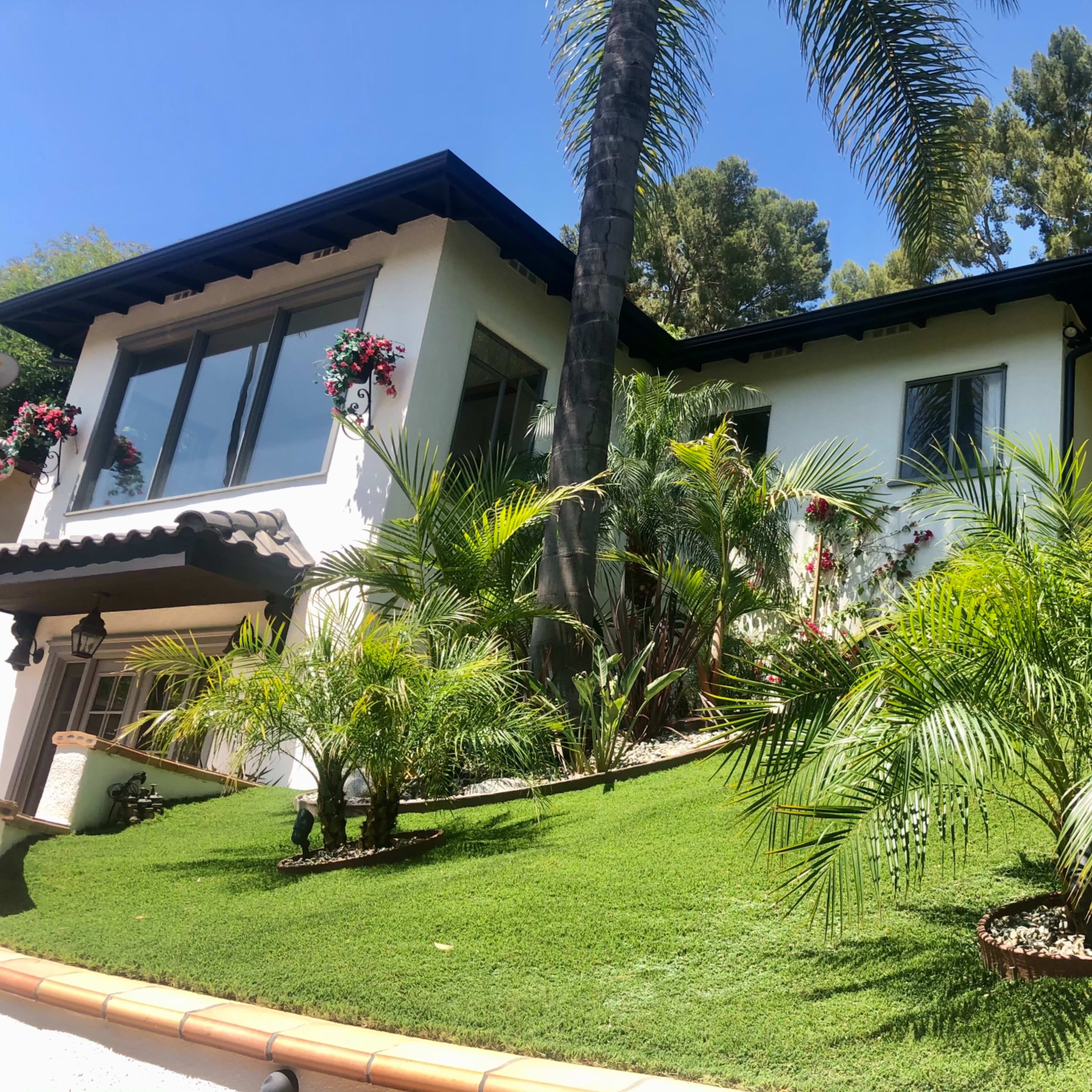 A two-story white house with a sloped green lawn, adorned with palm trees and flowering plants on the exterior.