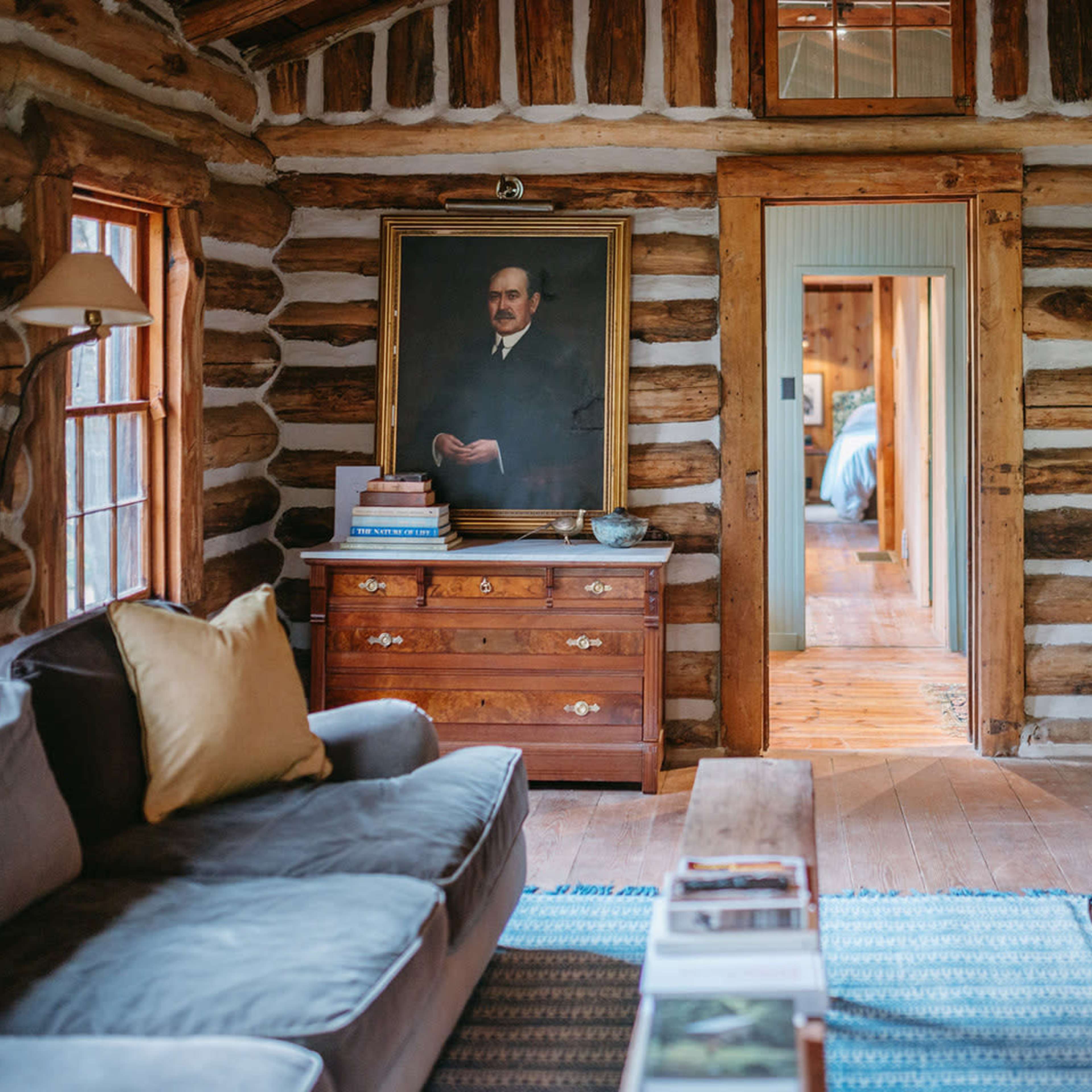 A cozy log cabin interior featuring a sofa, a wooden chest of drawers with a portrait above it, and a mounted deer head on the wall.