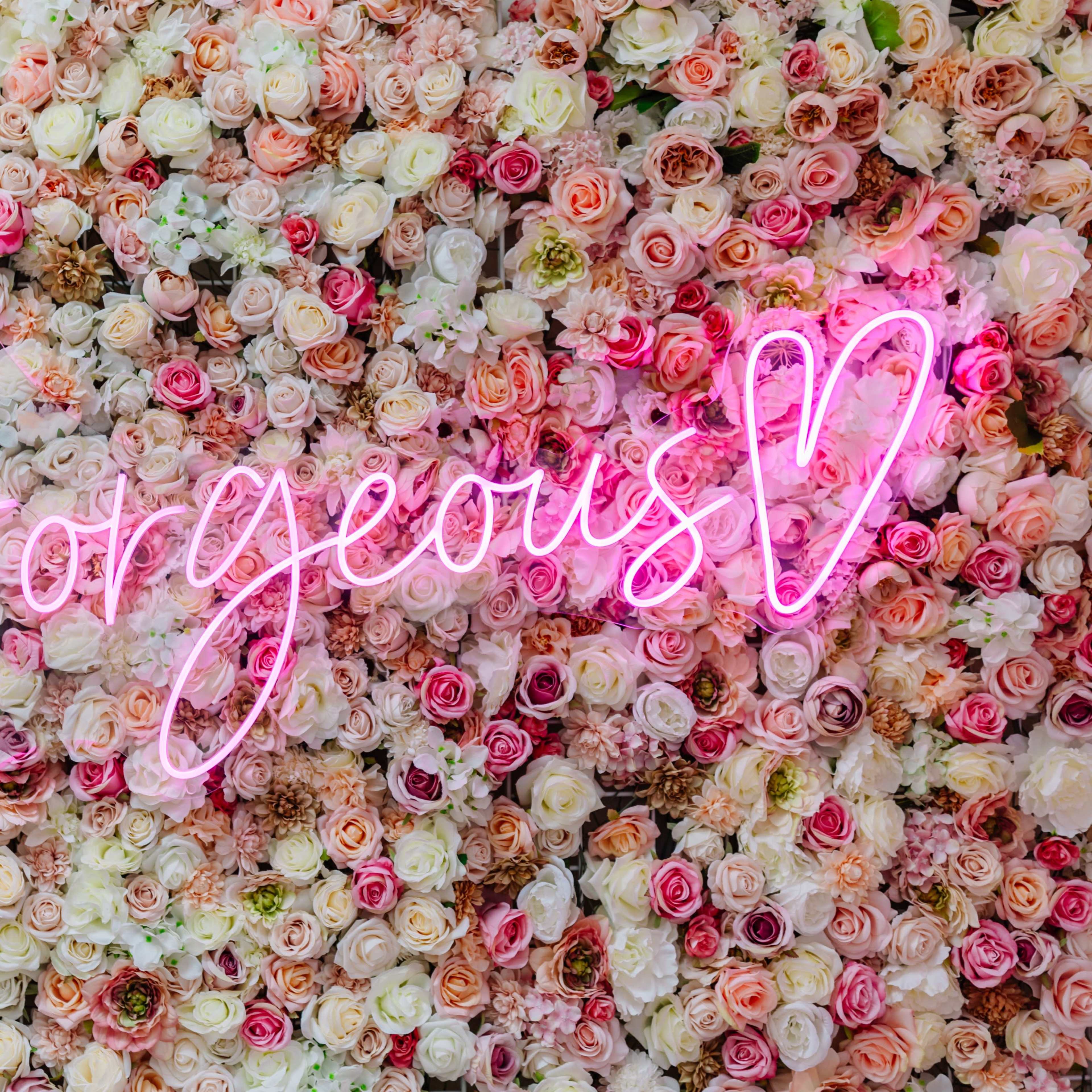 A wall covered in various colors of artificial roses features a pink neon sign that says "Gorgeous."