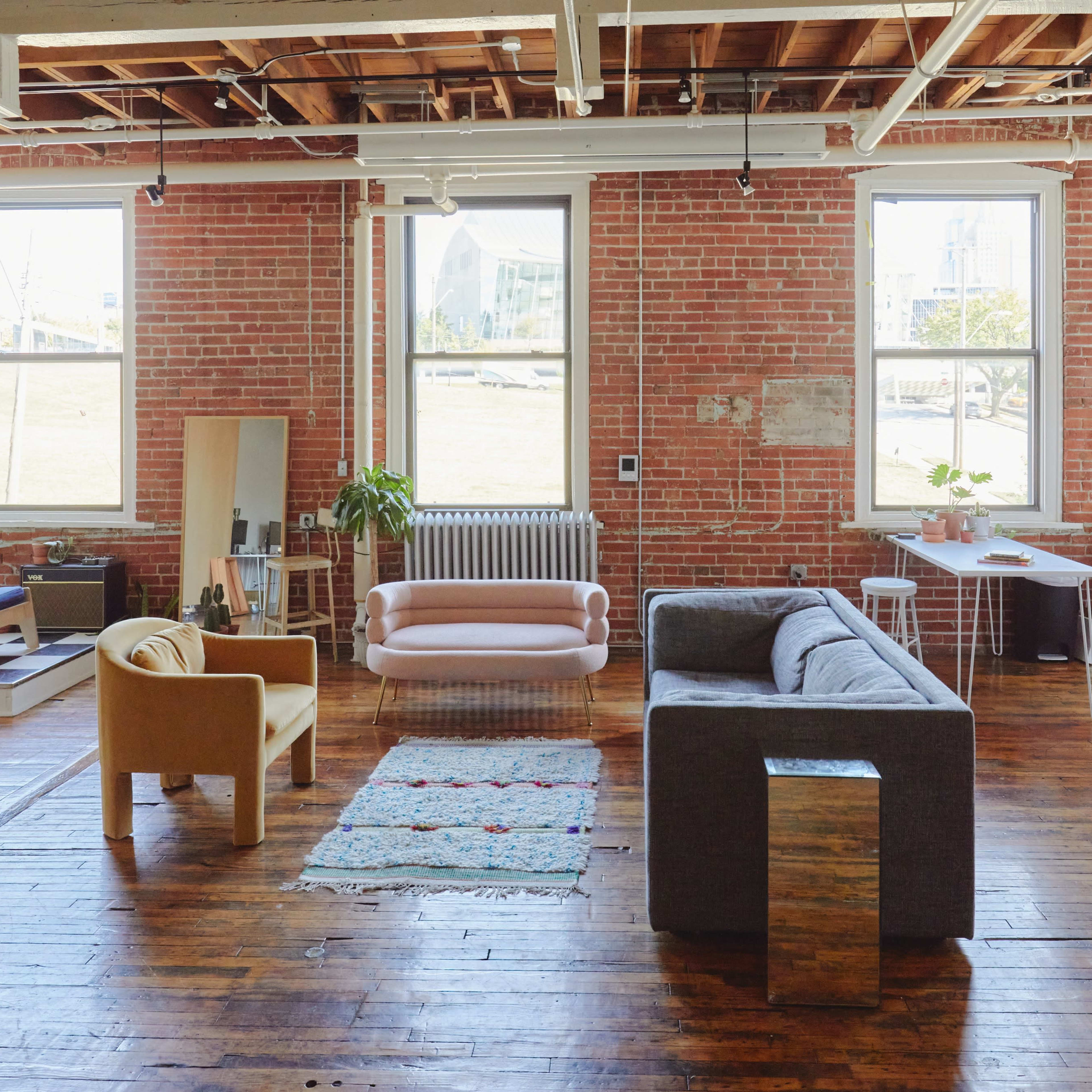 A spacious loft-style living area with exposed brick walls, hardwood floors, and various seating arrangements, including a gray sofa and a pink chair, along with plants and windows allowing natural light.