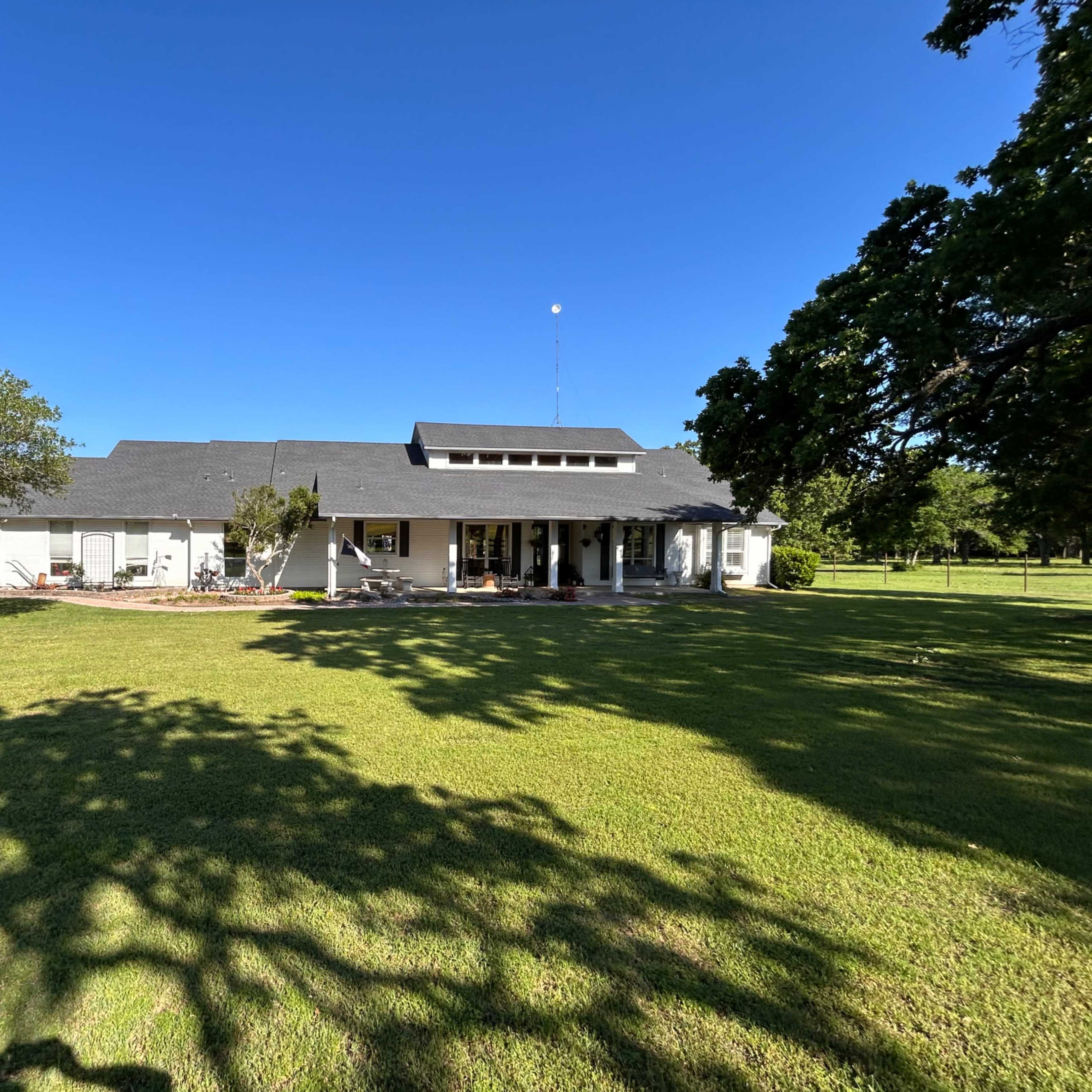 A single-story house with a grey roof and front porch is set on a green lawn surrounded by trees and a gravel driveway.