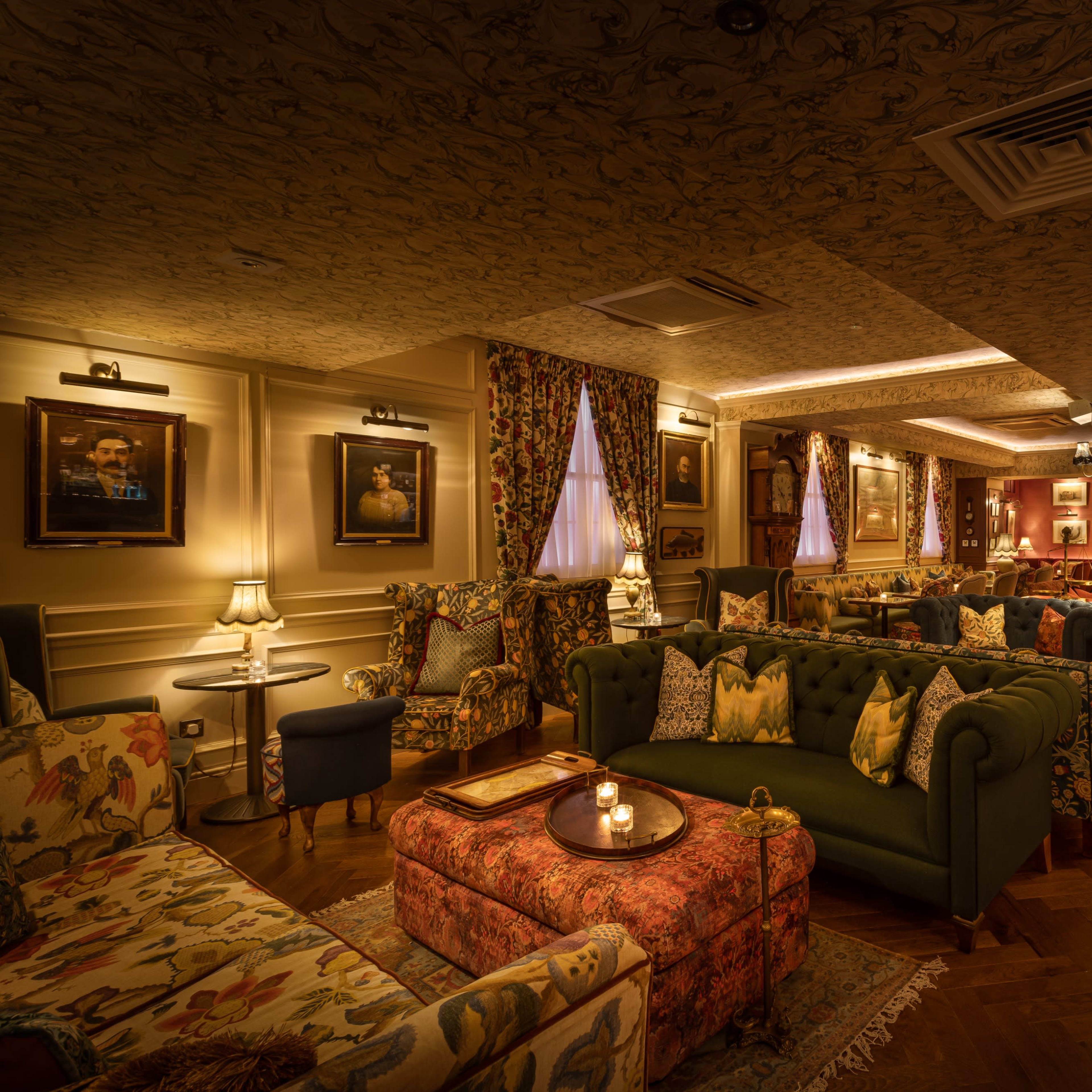 The interior of a cozy lounge features patterned sofas, floral curtains, and framed artwork on the walls, with warm lighting creating an inviting atmosphere.
