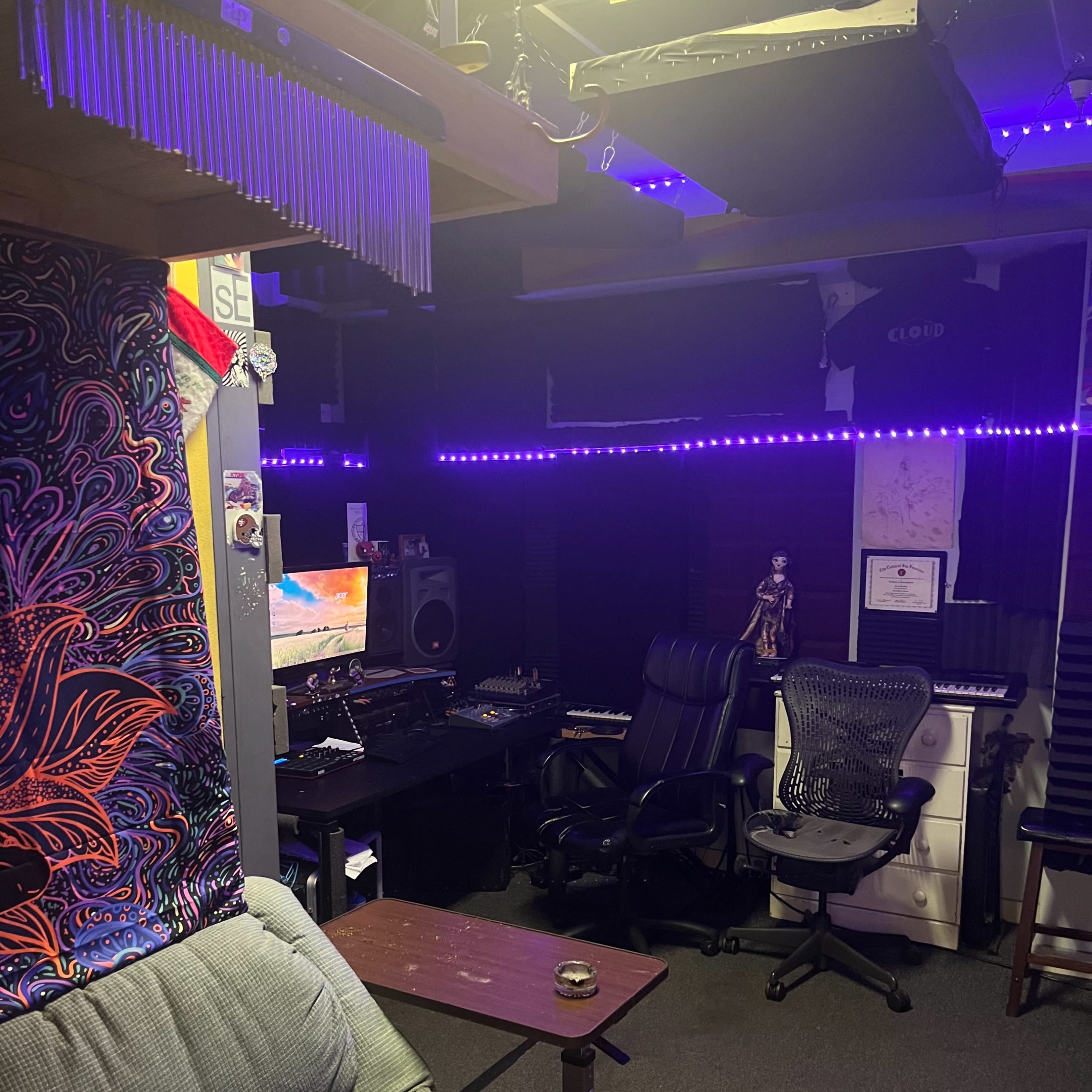 A music production studio with a couch, two office chairs, a computer desk, and colorful LED lighting.
