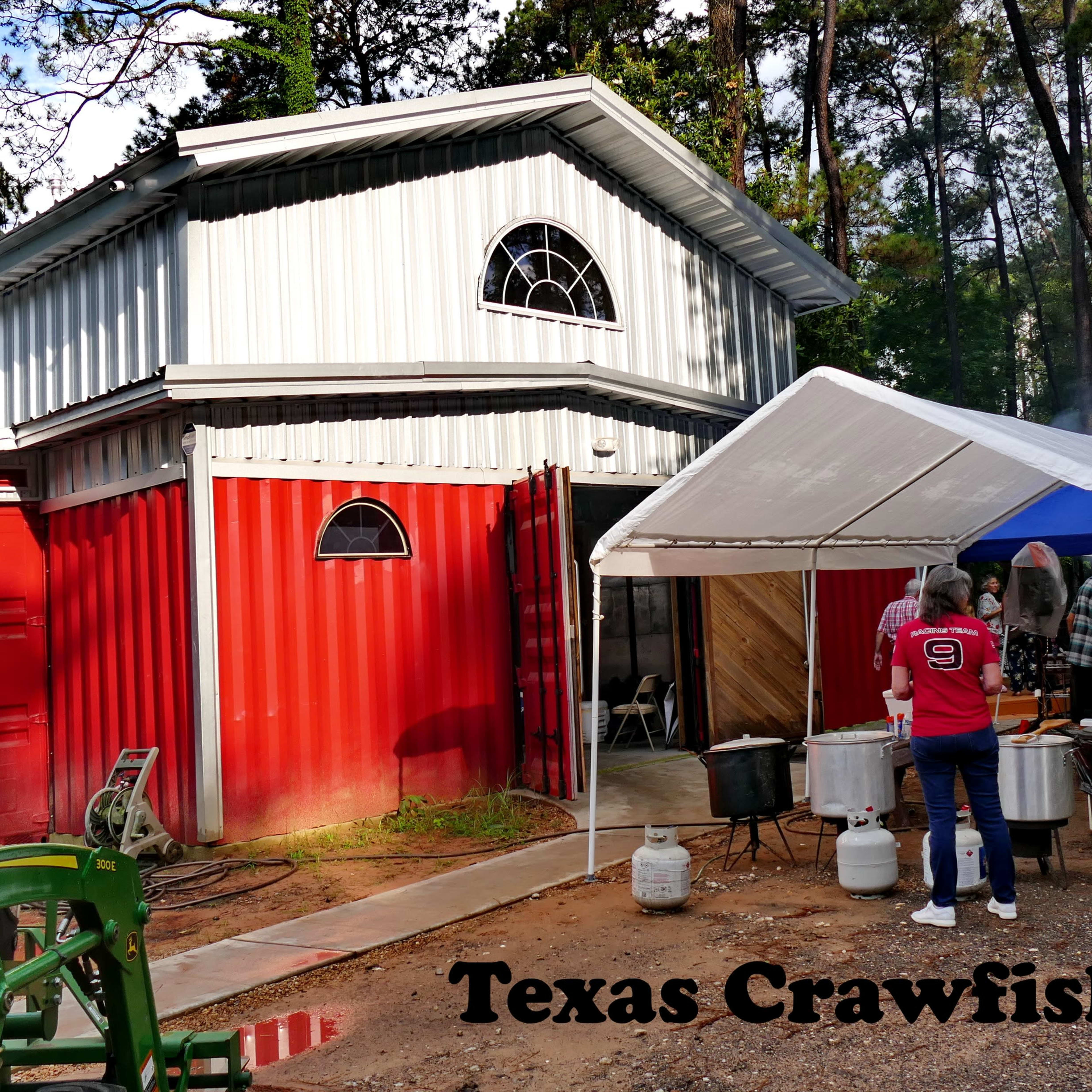 A person stands under a tent near a barn with red and silver exterior, preparing for a crawfish boil in a wooded area.