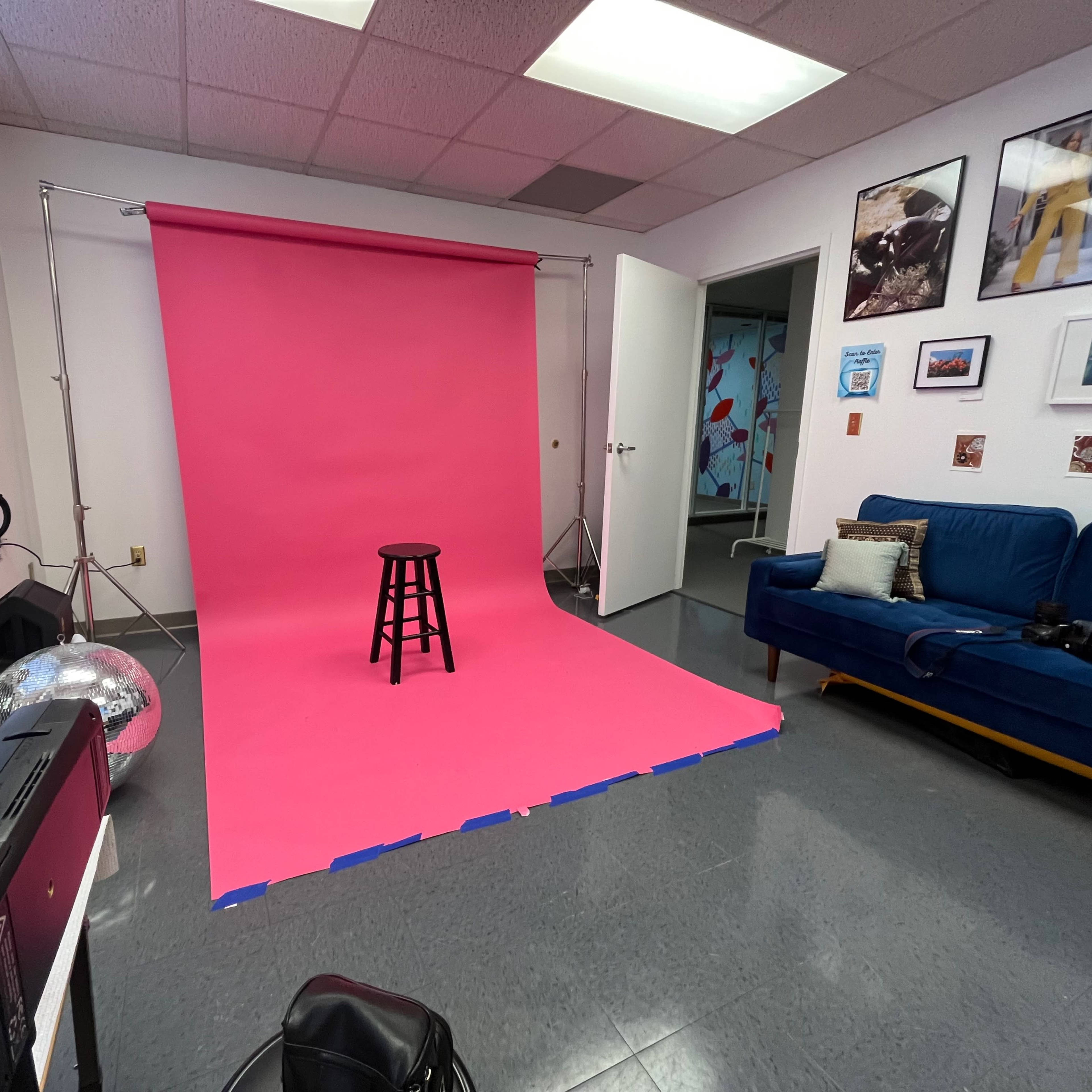 A pink backdrop with a black stool set up in a photography studio, alongside a blue couch and various framed pictures on the walls.