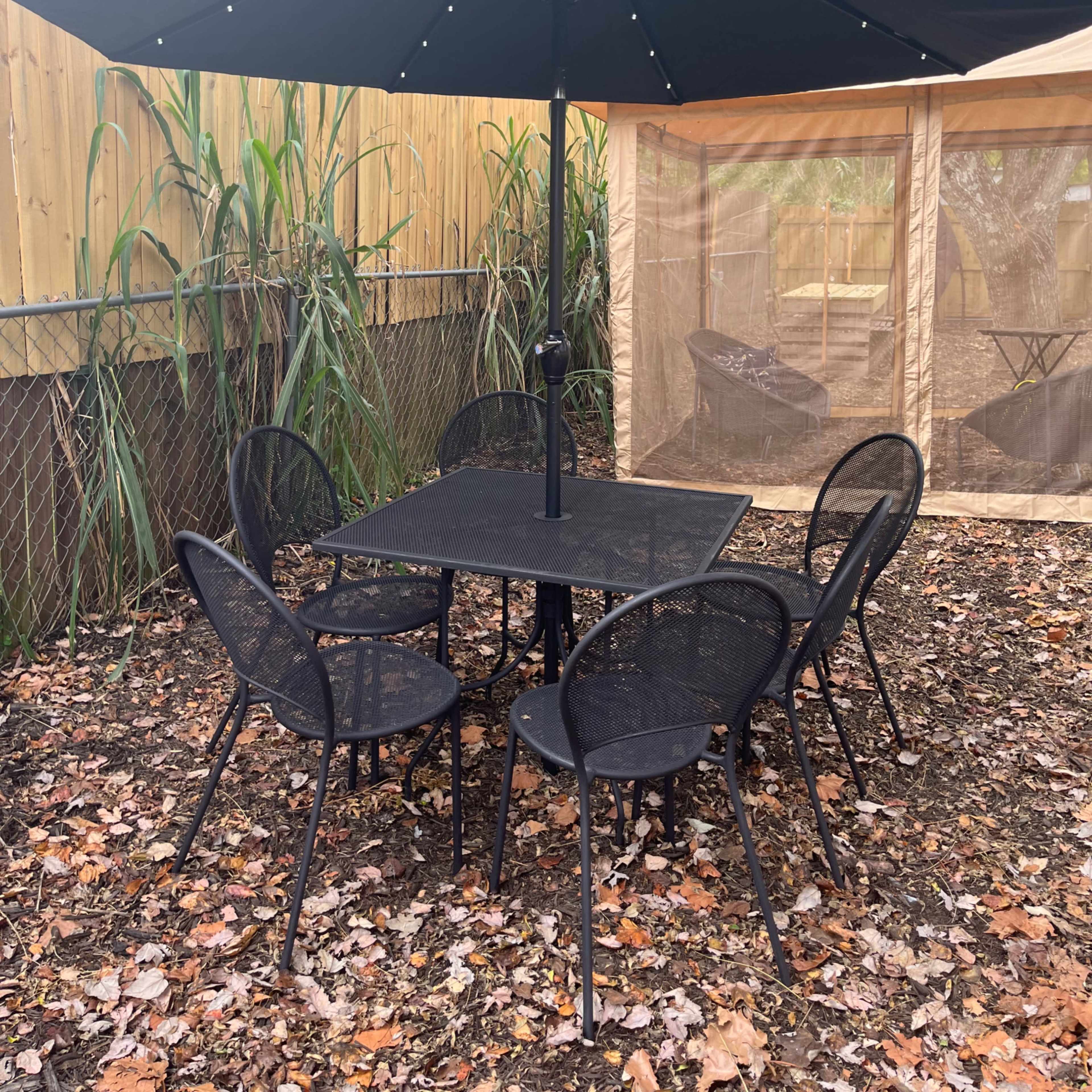 A black metal table with six matching chairs is set beneath a large black umbrella in a backyard surrounded by greenery and a tan tent.