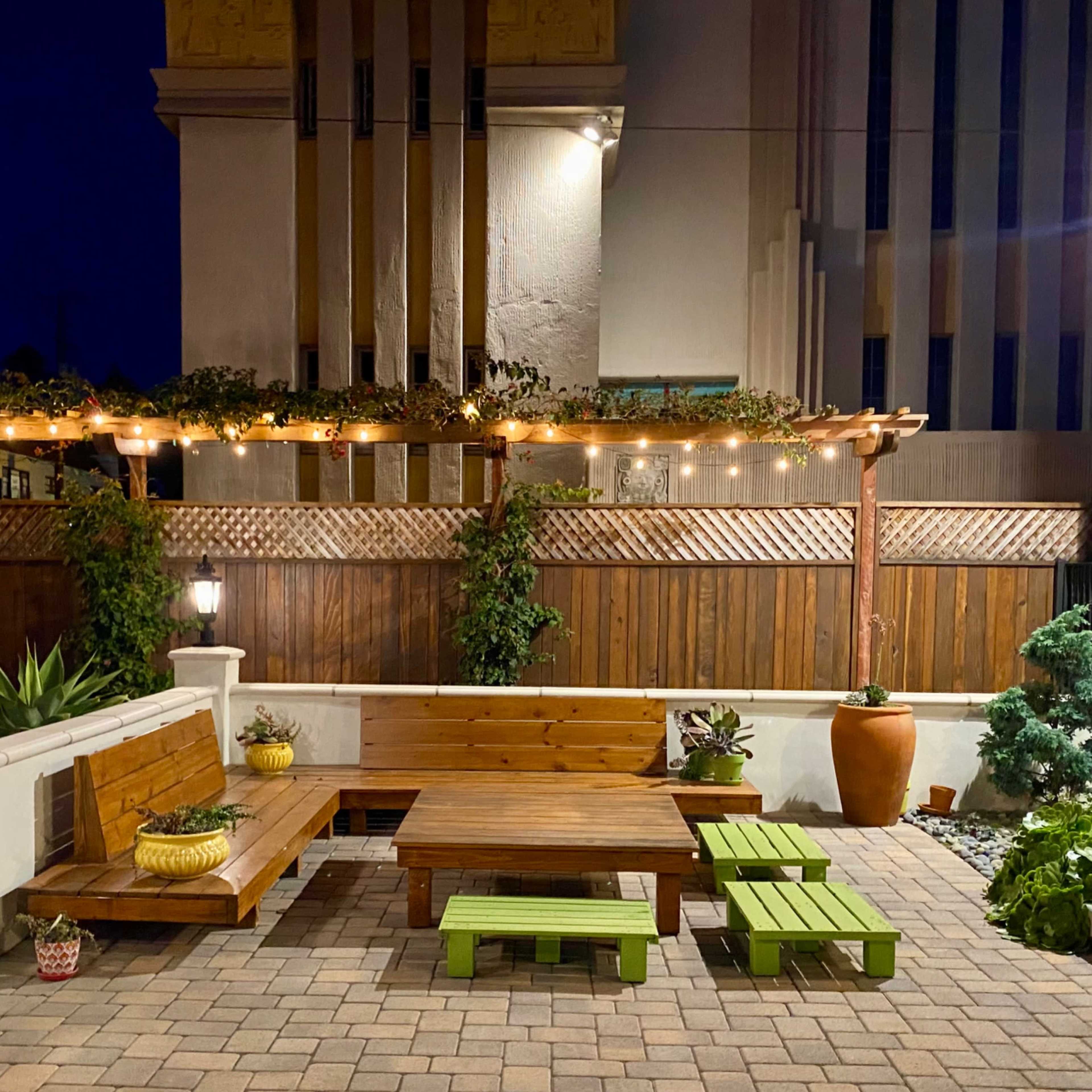 A well-lit outdoor patio area featuring wooden benches, green tables, and string lights overhead against a backdrop of a building with vertical stripes.