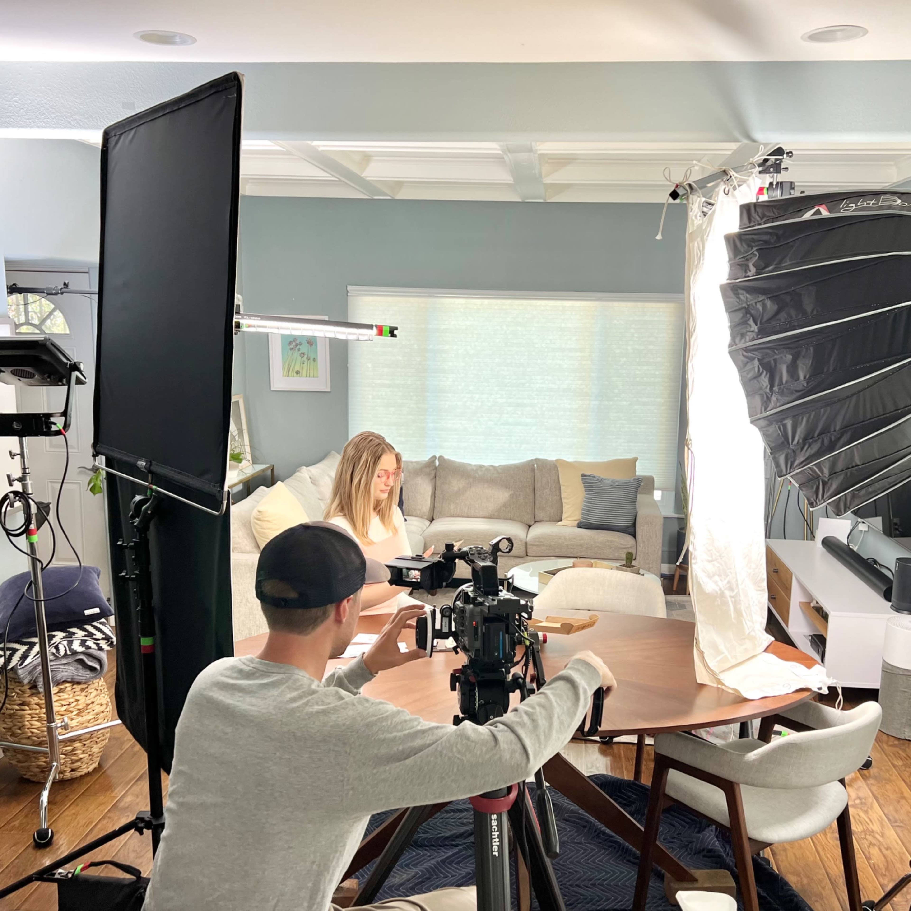 A person sits at a table in a living room while a crew sets up video lighting and camera equipment around them.
