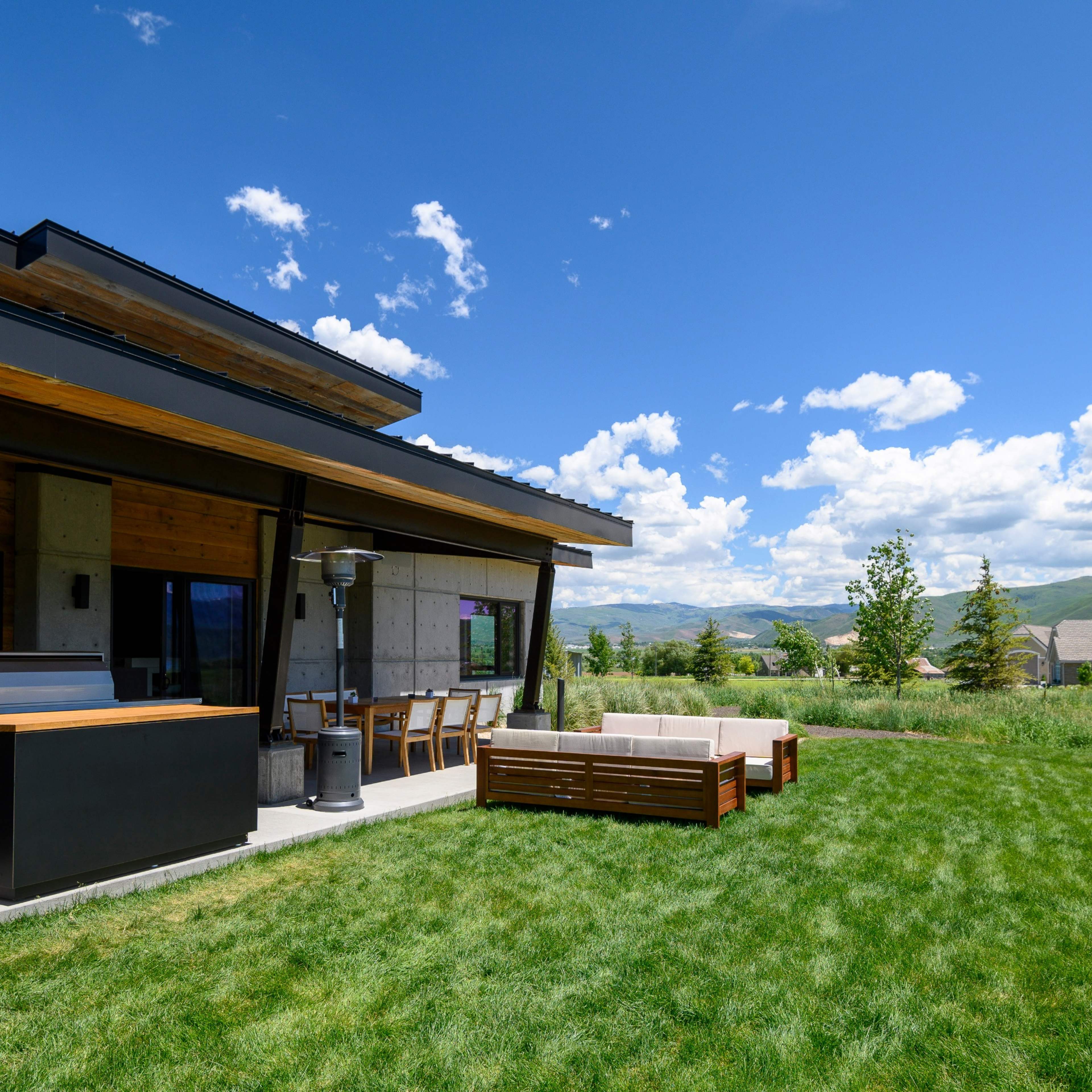 A modern house with a patio area featuring outdoor seating and a grill overlooks a green grassy landscape under a blue sky with clouds.
