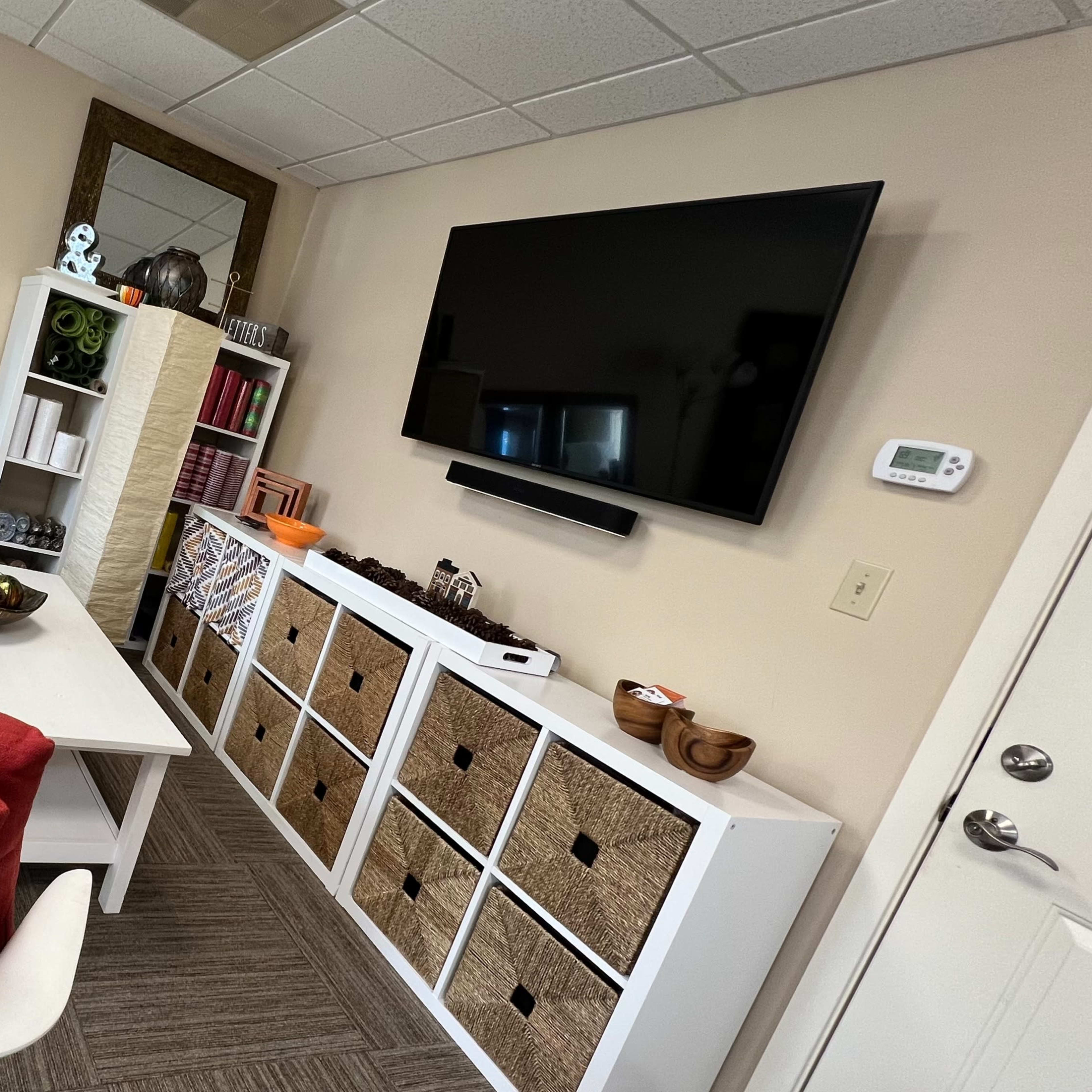 A modern office or lounge area with a white sofa, a white coffee table, storage cubbies, a wall-mounted television, and decorative items.