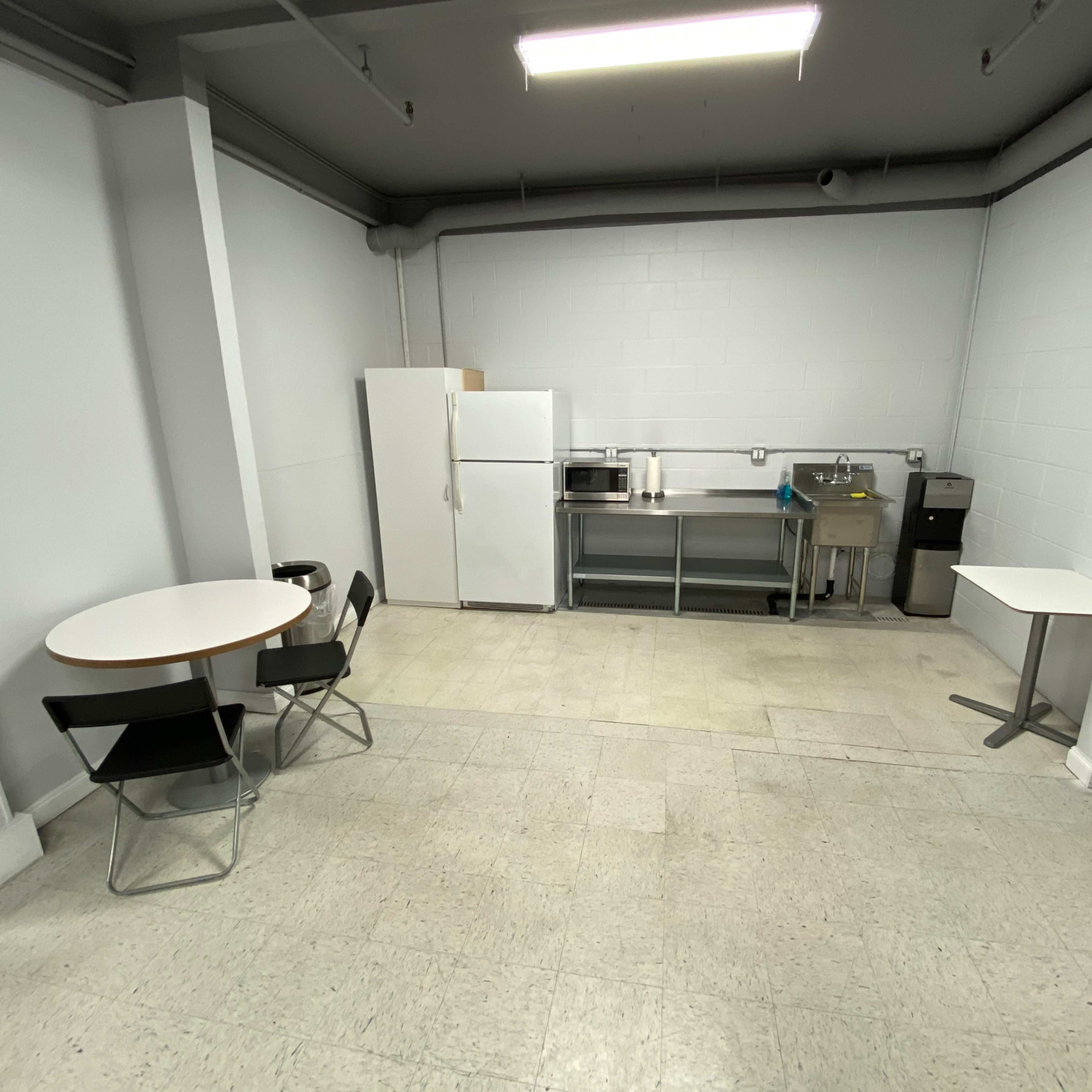 A sparse kitchen area featuring a white refrigerator, a microwave, a sink, and two tables with chairs arranged in a corner.