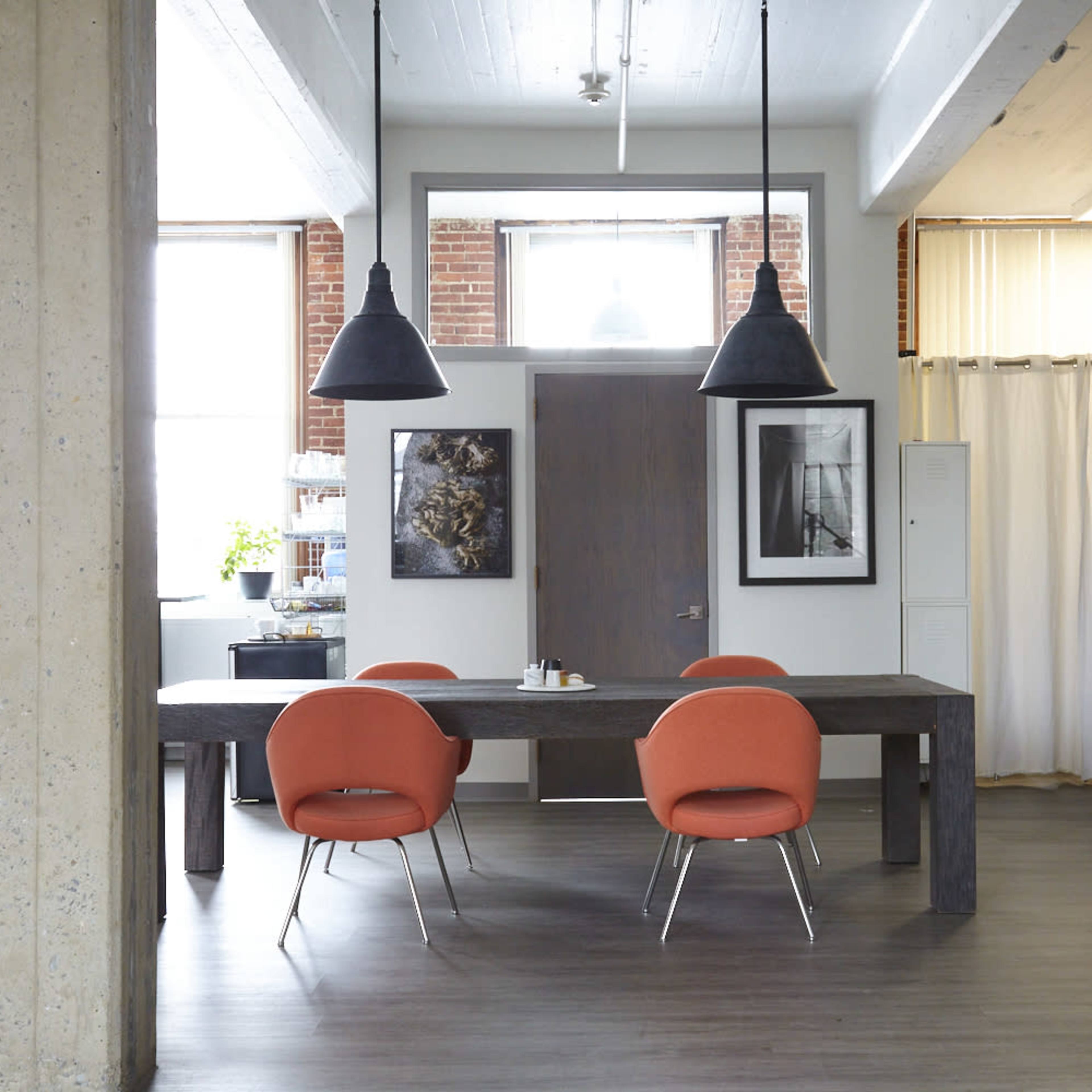 A modern dining area featuring a large wooden table with four orange chairs, two pendant lights hanging from the ceiling, and large windows allowing natural light to enter.