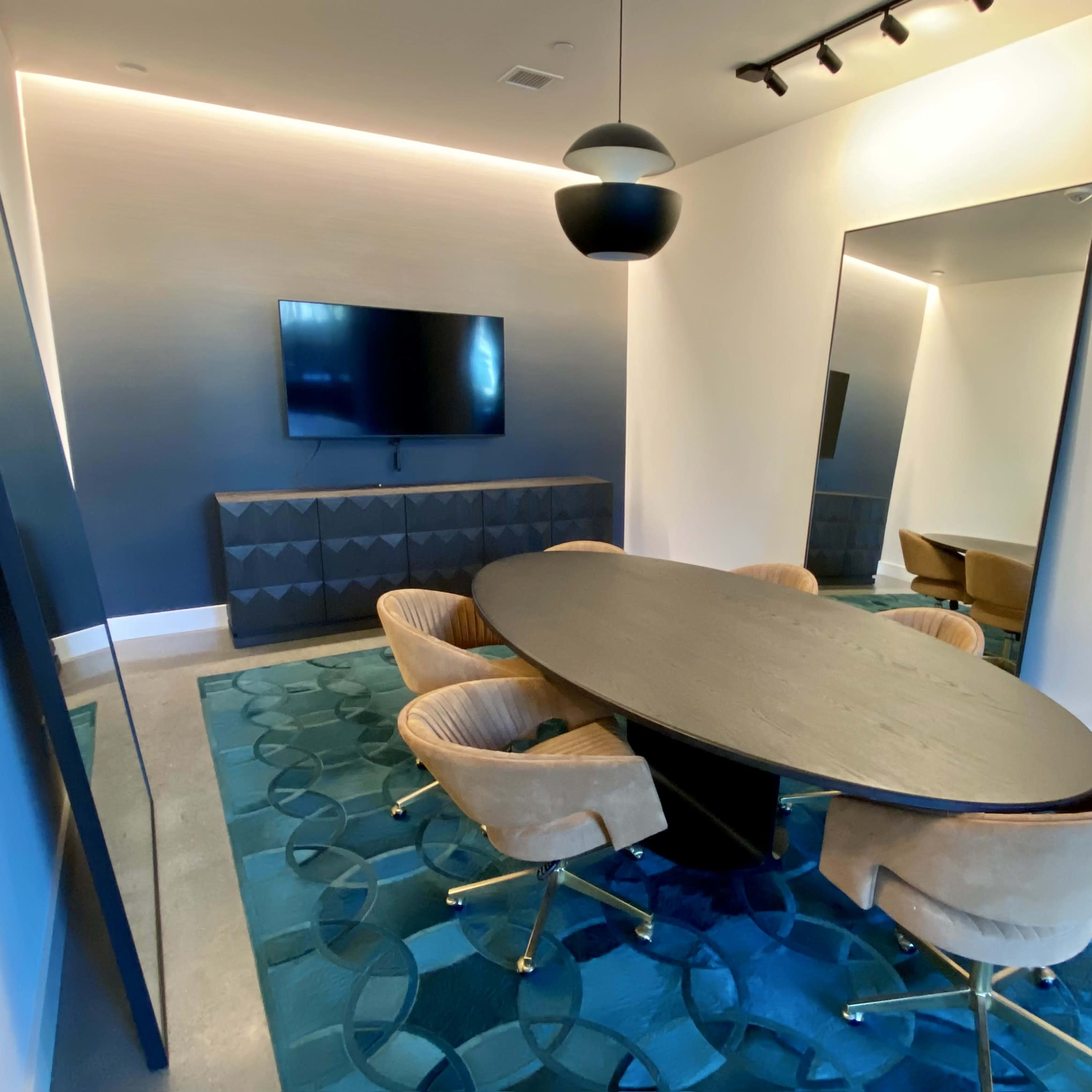 A modern conference room features an oval table surrounded by four upholstered chairs, a wall-mounted television, and a large mirror reflecting the space.