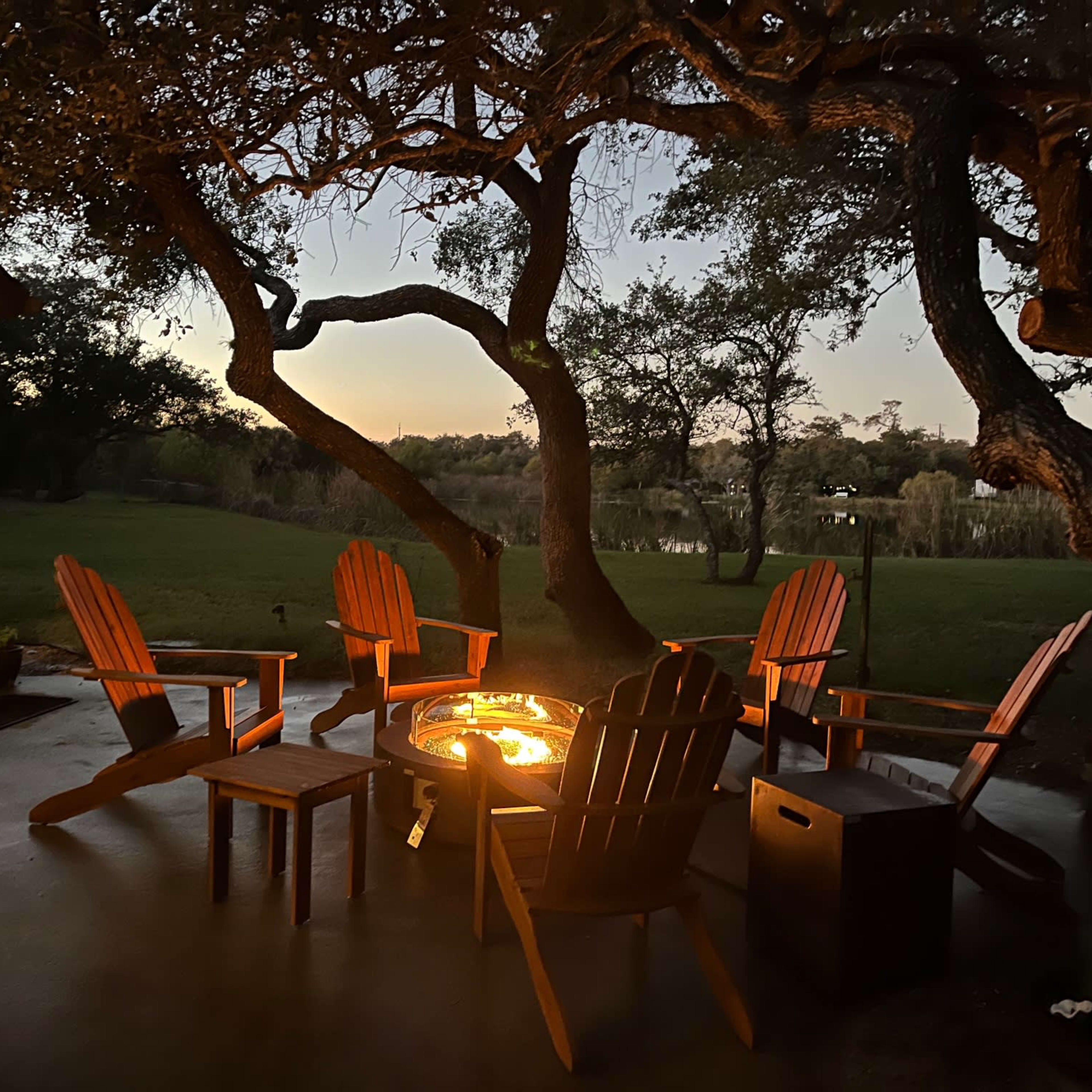 A circle of wooden chairs surrounds a fire pit under large trees at dusk, overlooking a grassy area and water in the background.