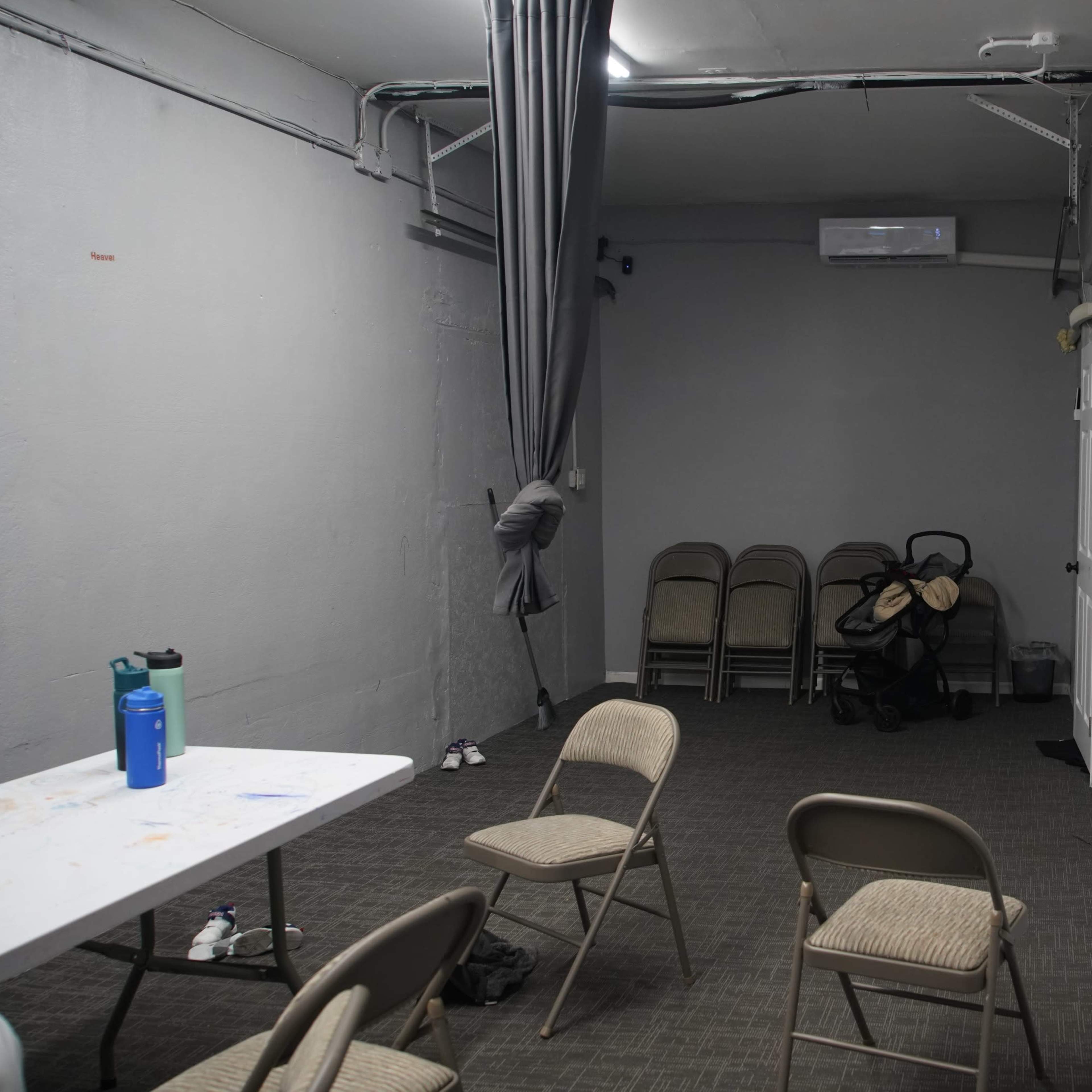 A small, empty room with gray walls, a table, several folding chairs, and a stroller in the corner.