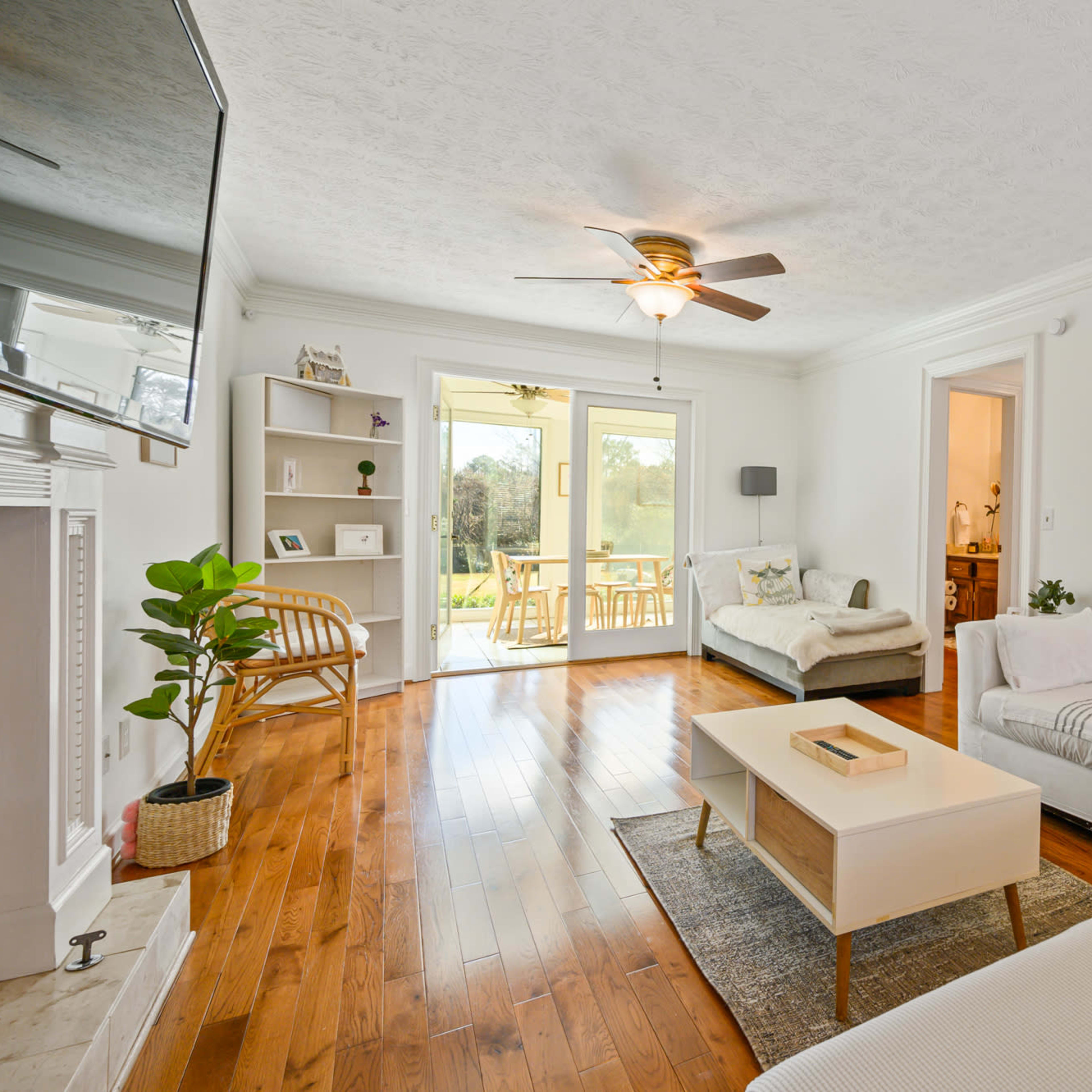 A bright living room with a wooden floor, a ceiling fan, a white sectional sofa, and a coffee table, while large doors lead to a patio area.
