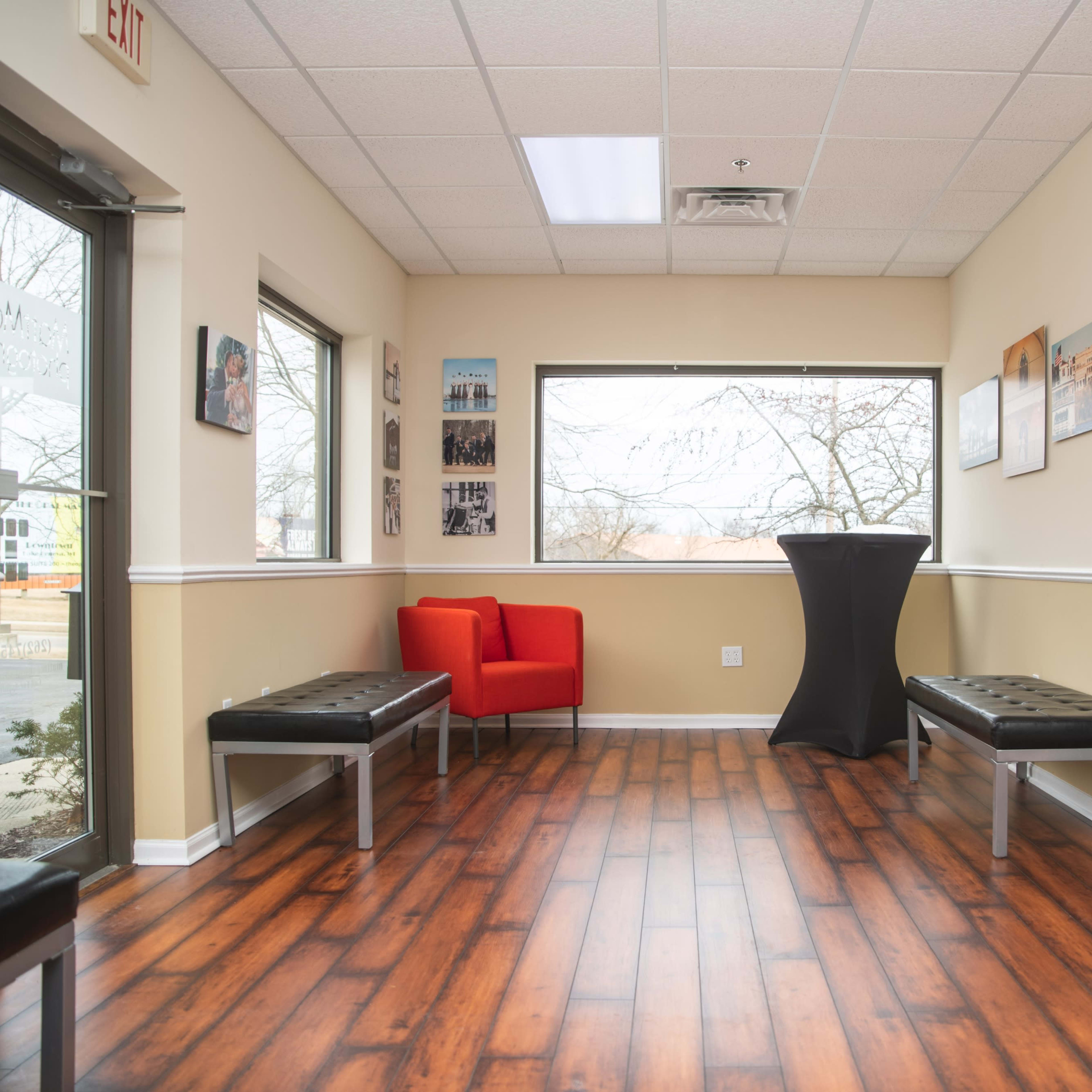A waiting room with wooden flooring, two benches, a red chair, and a tall black table near a large window.