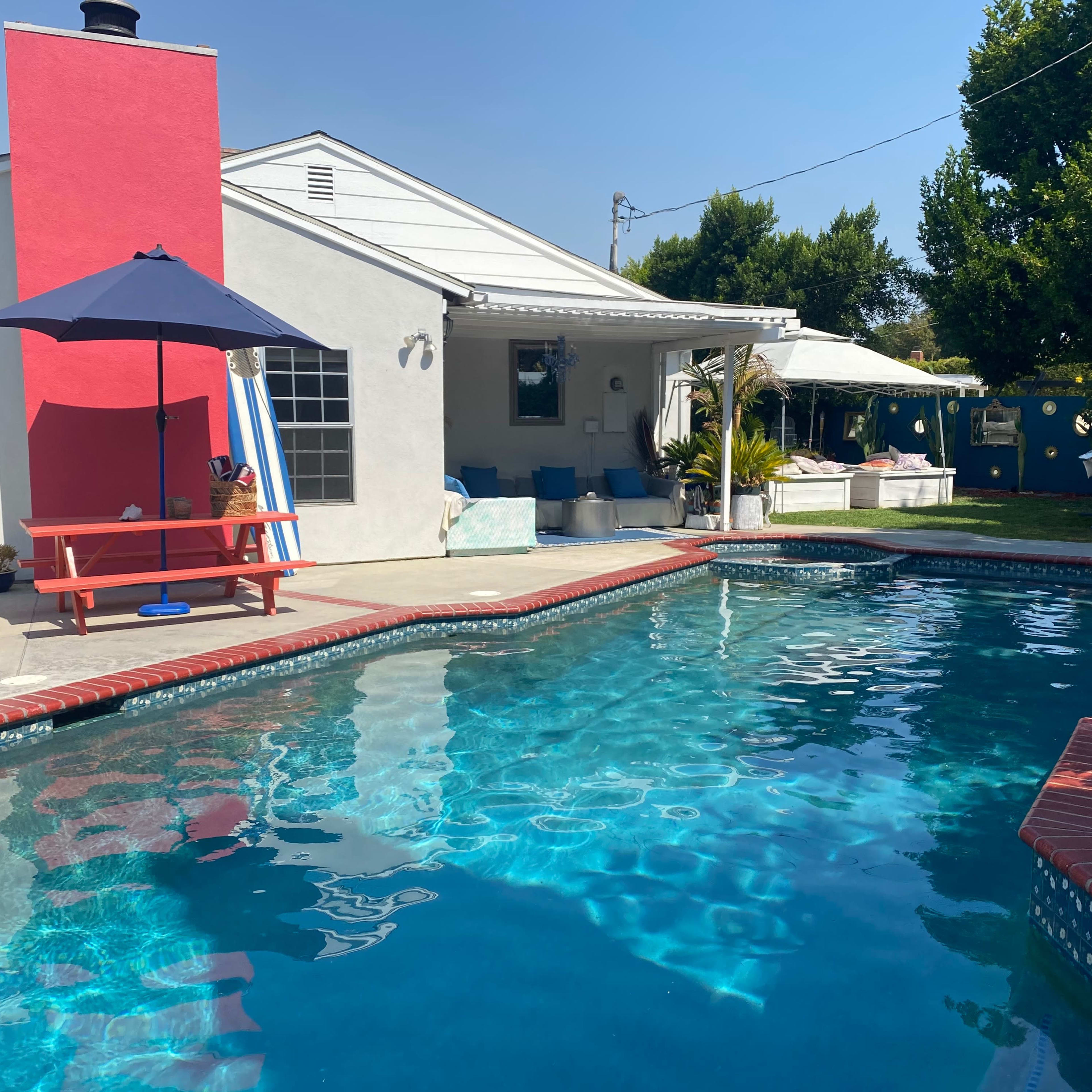 A clear blue swimming pool is surrounded by a patio with a red table and umbrella next to a house featuring a bright red accent wall.
