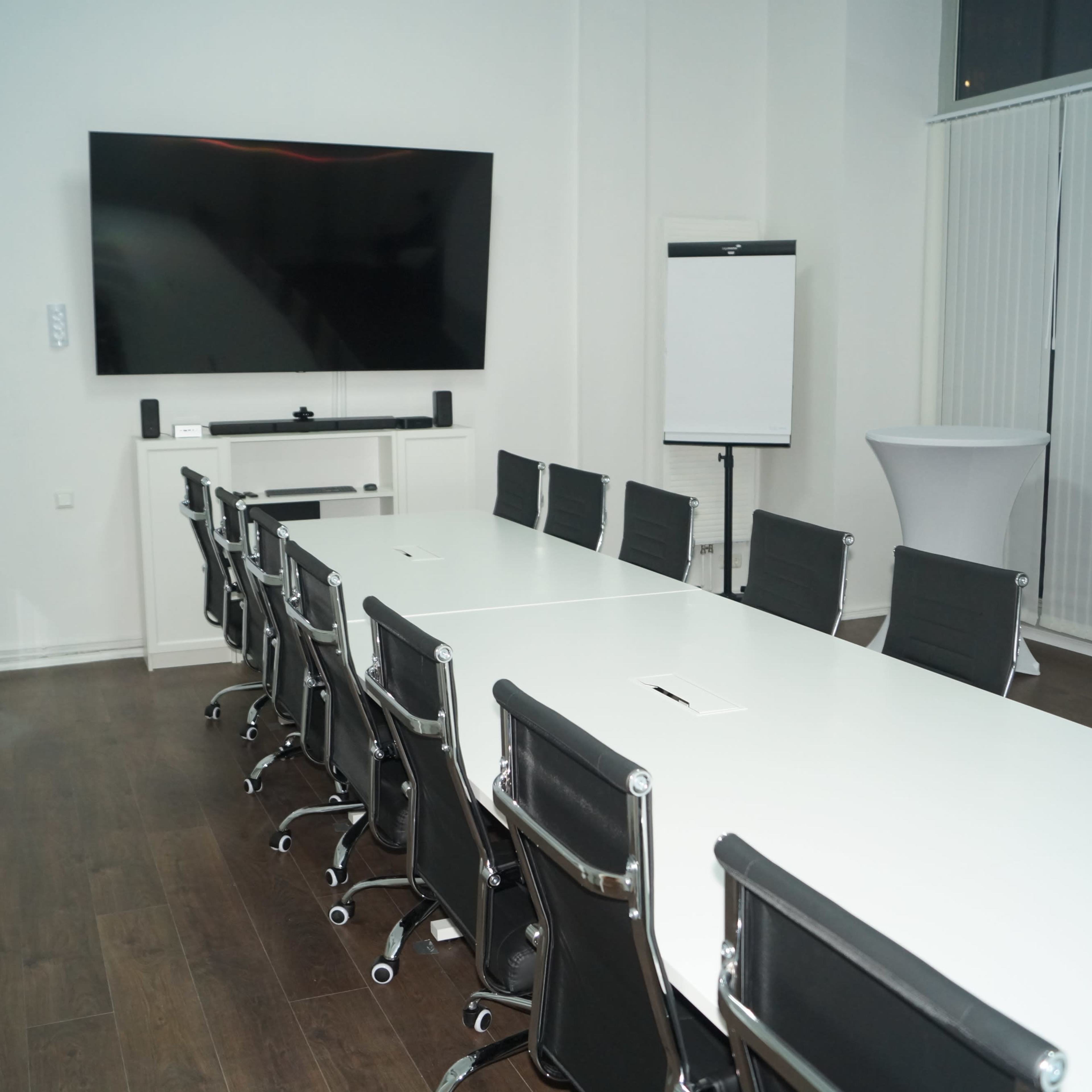 A modern conference room features a long white table with black rolling chairs and a large screen mounted on the wall.