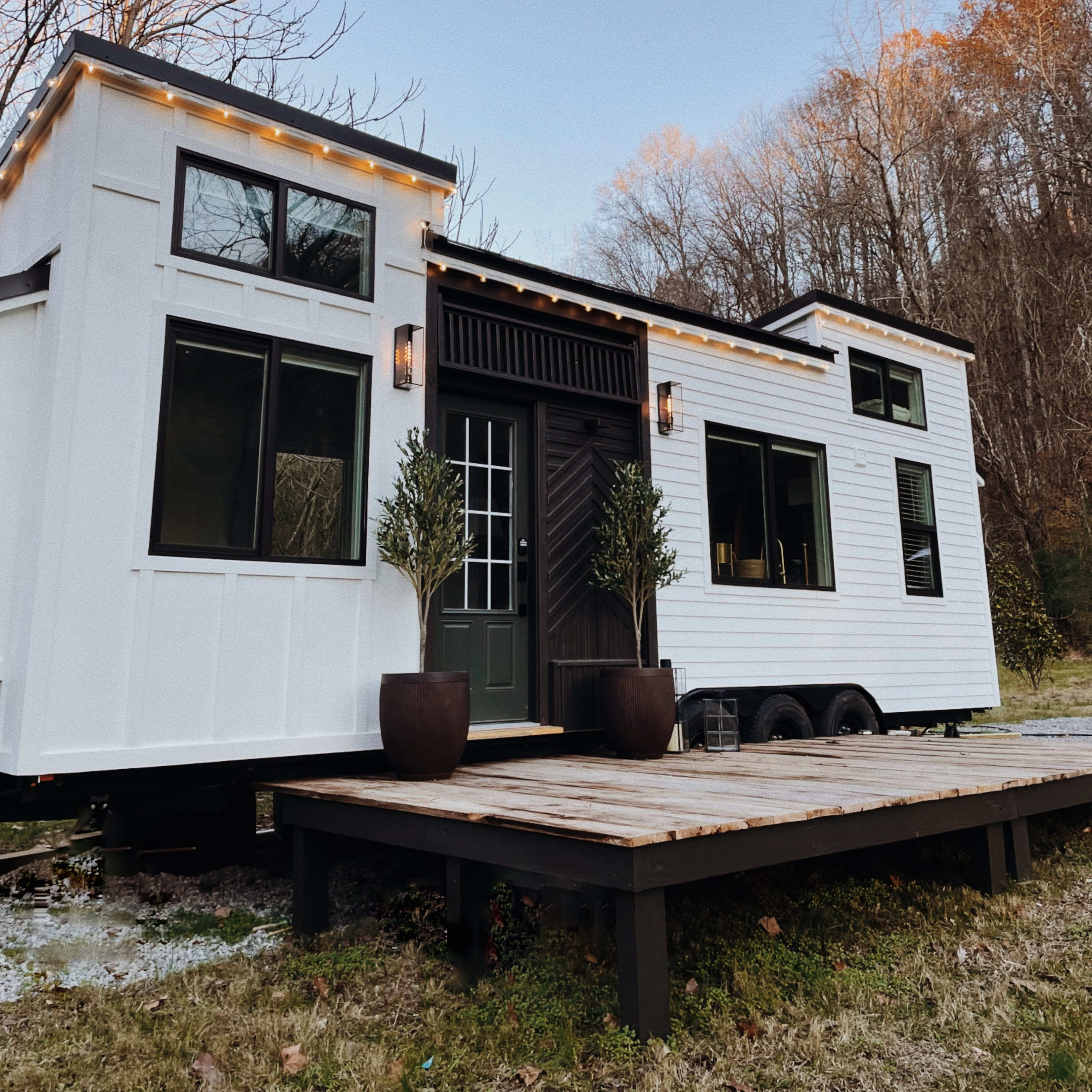 A modern tiny house on wheels with a wood deck, surrounded by trees and a clear sky.