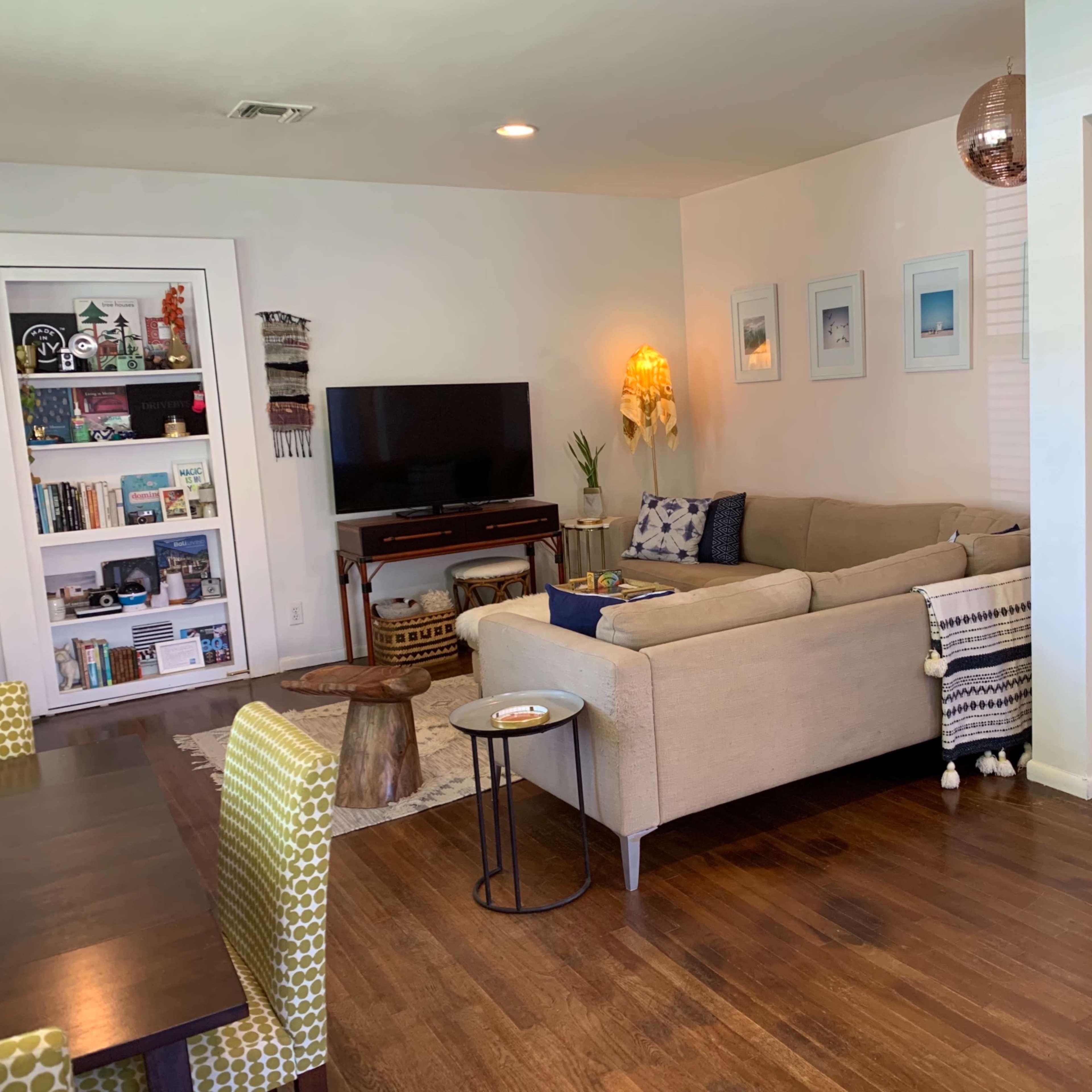 A modern living room with a beige sectional sofa, a television on a stand, a bookshelf filled with books, and a decorative mirror on the wall.