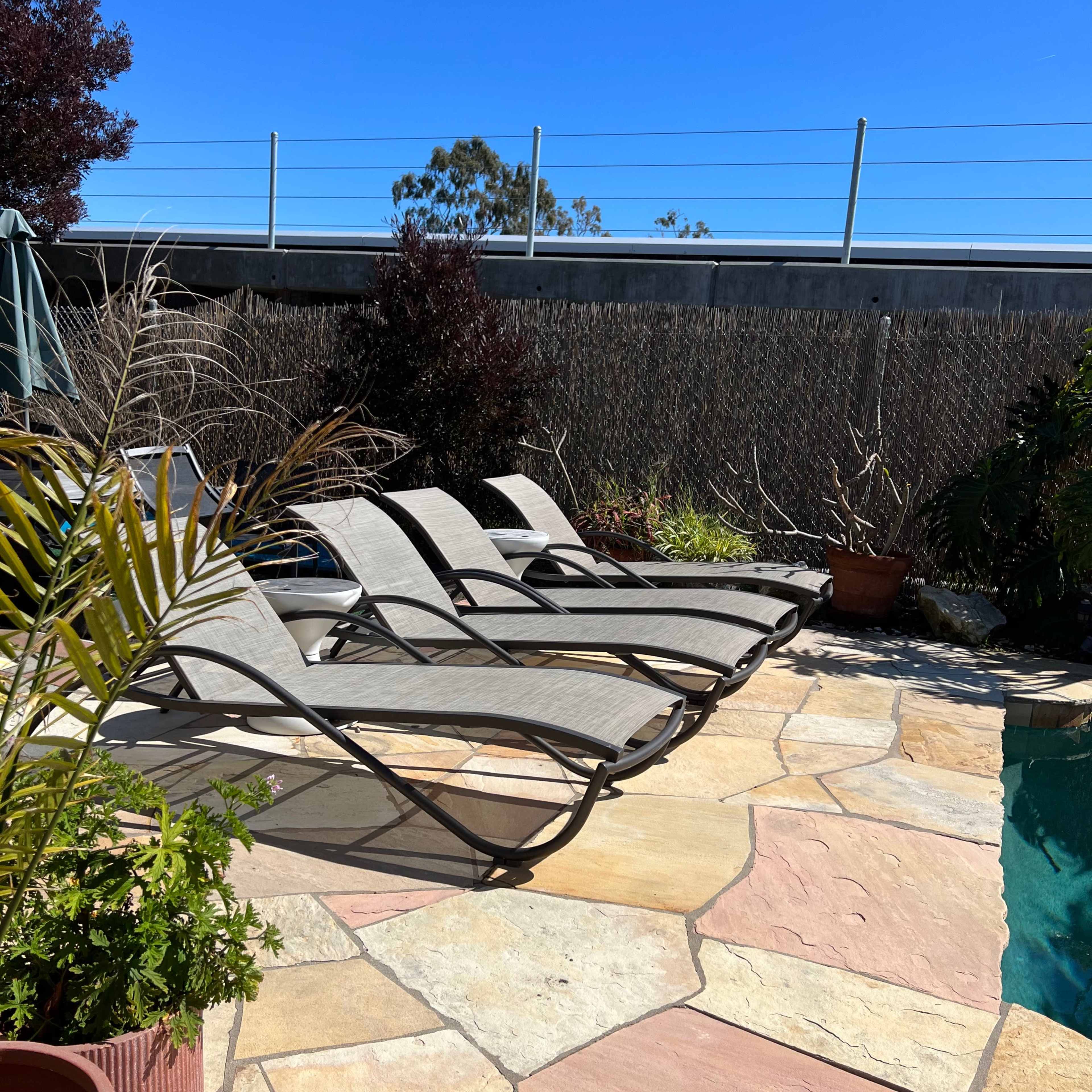 A row of lounge chairs is arranged beside a stone patio near a swimming pool, surrounded by potted plants and a clear blue sky.