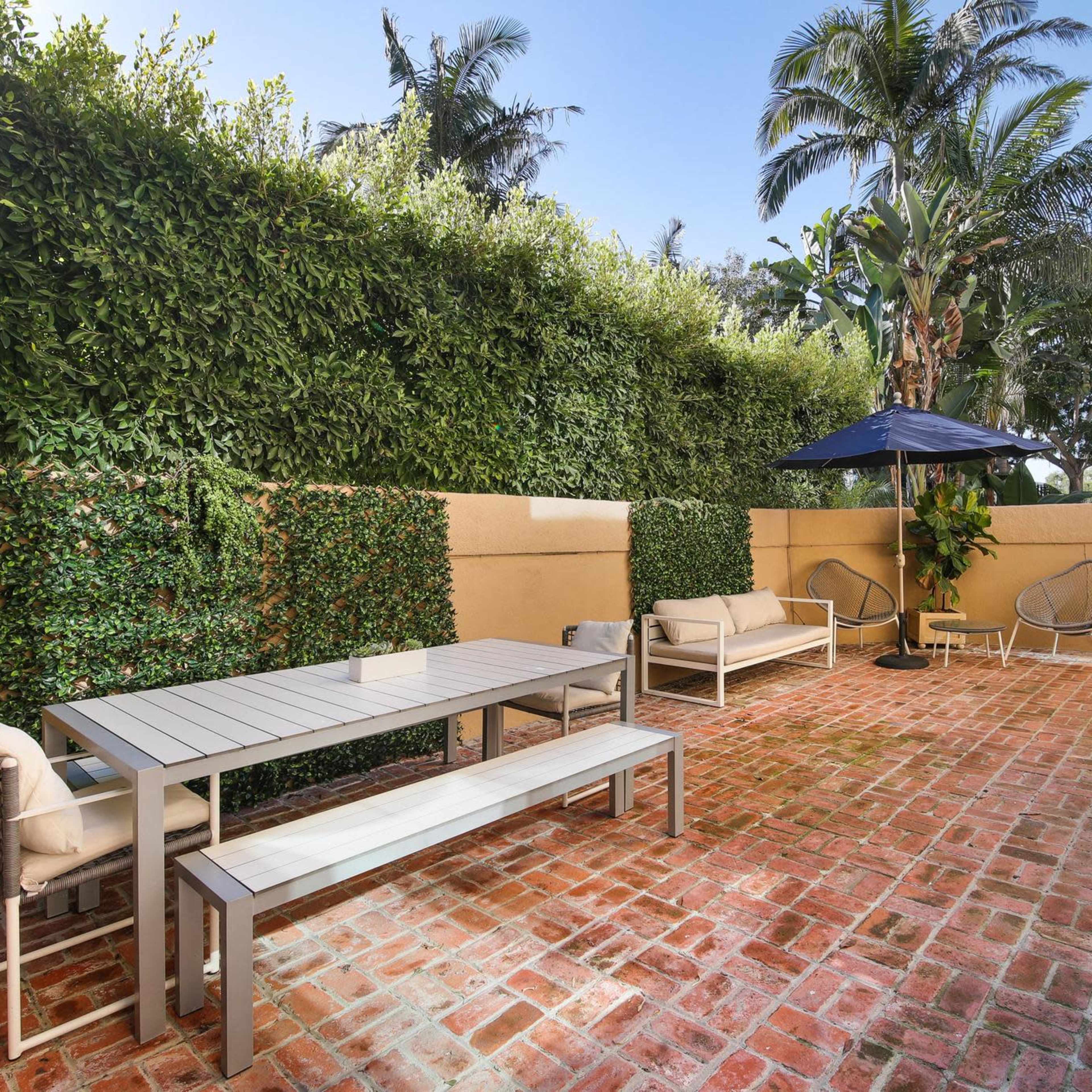 A patio area with a brick floor, a dining table, seating arrangements, and a large umbrella, surrounded by lush greenery and palm trees.