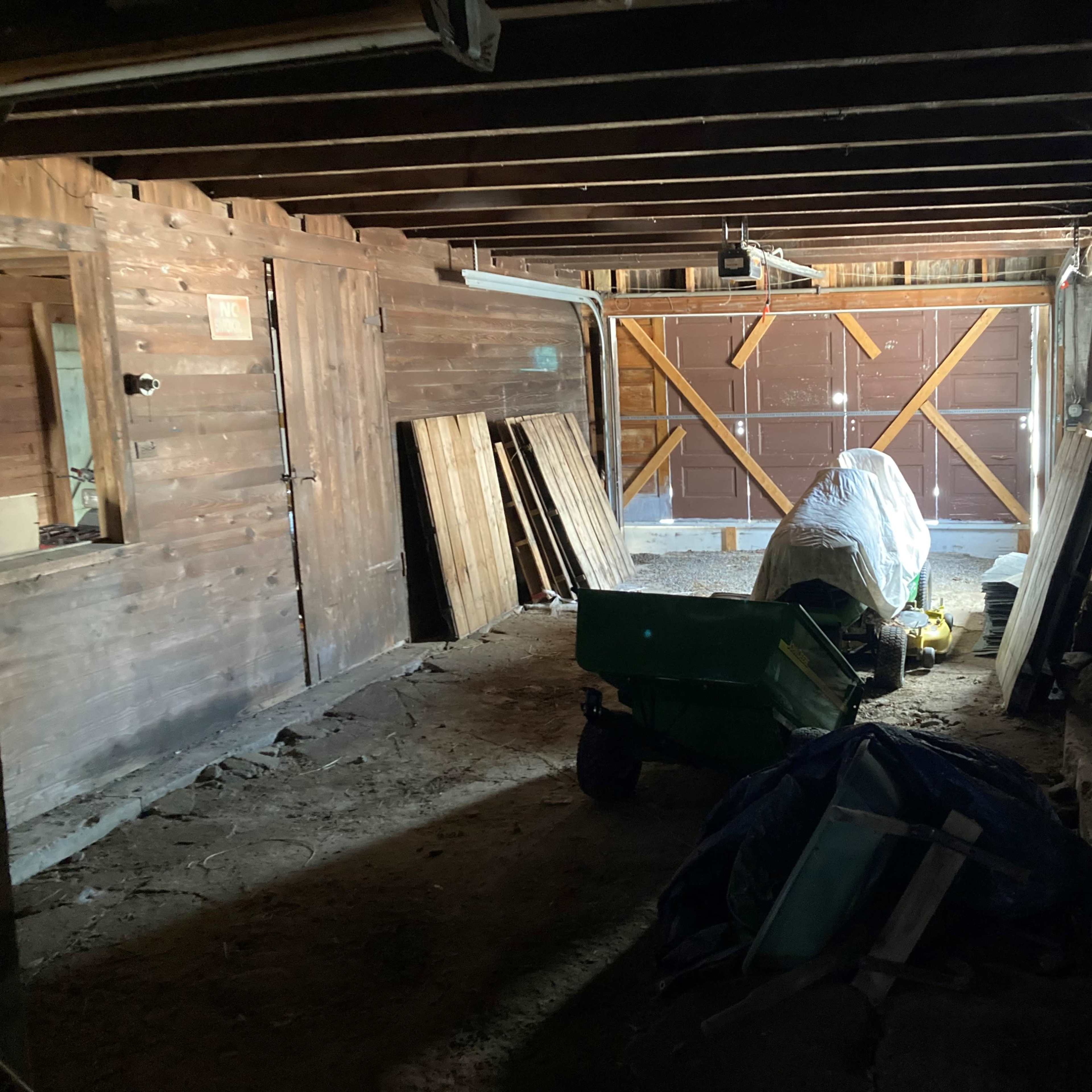 The interior of a dimly lit barn features wooden beams, storage materials against the walls, and a covered vehicle at the center.