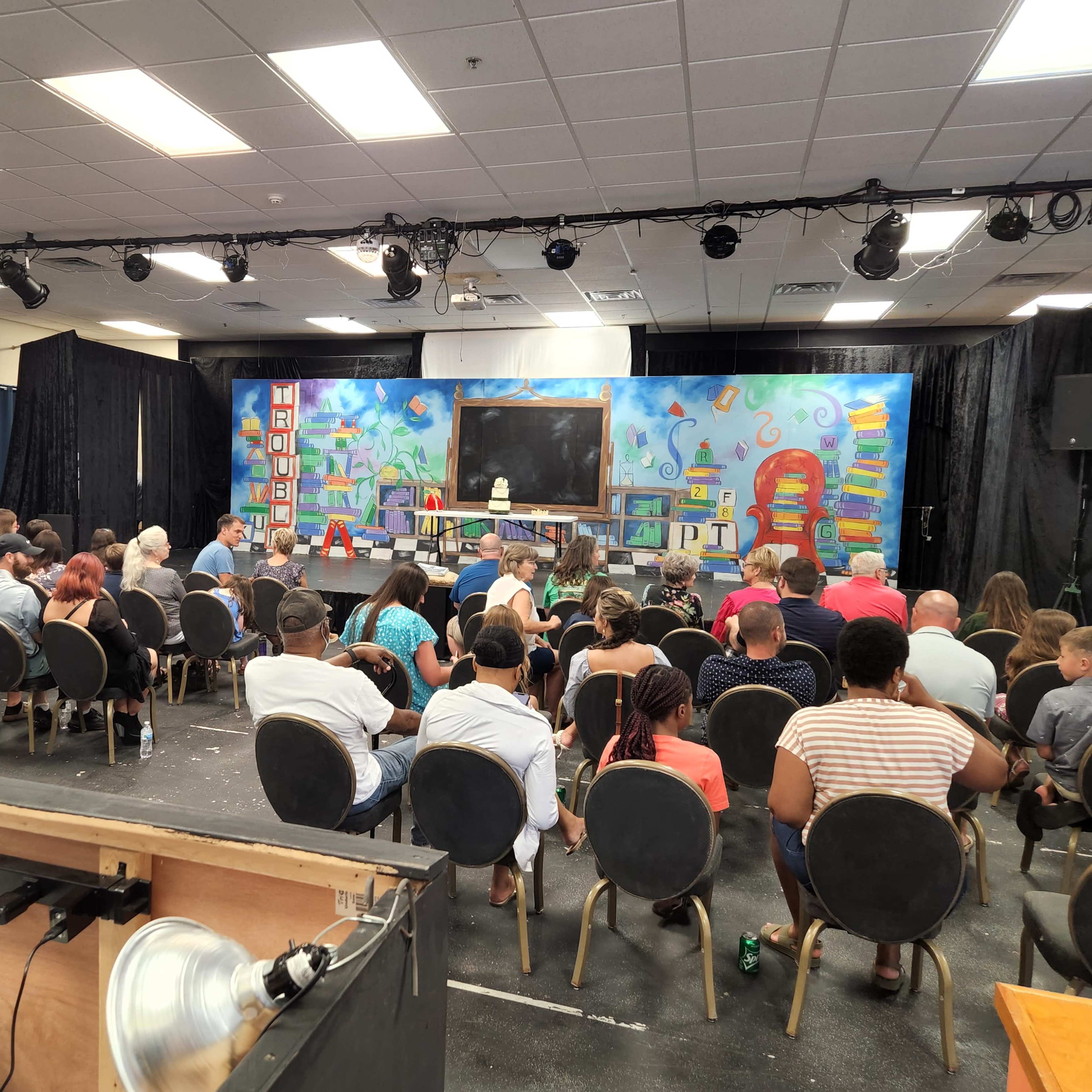 A group of people sits in rows facing a stage adorned with a colorful backdrop featuring various educational elements.