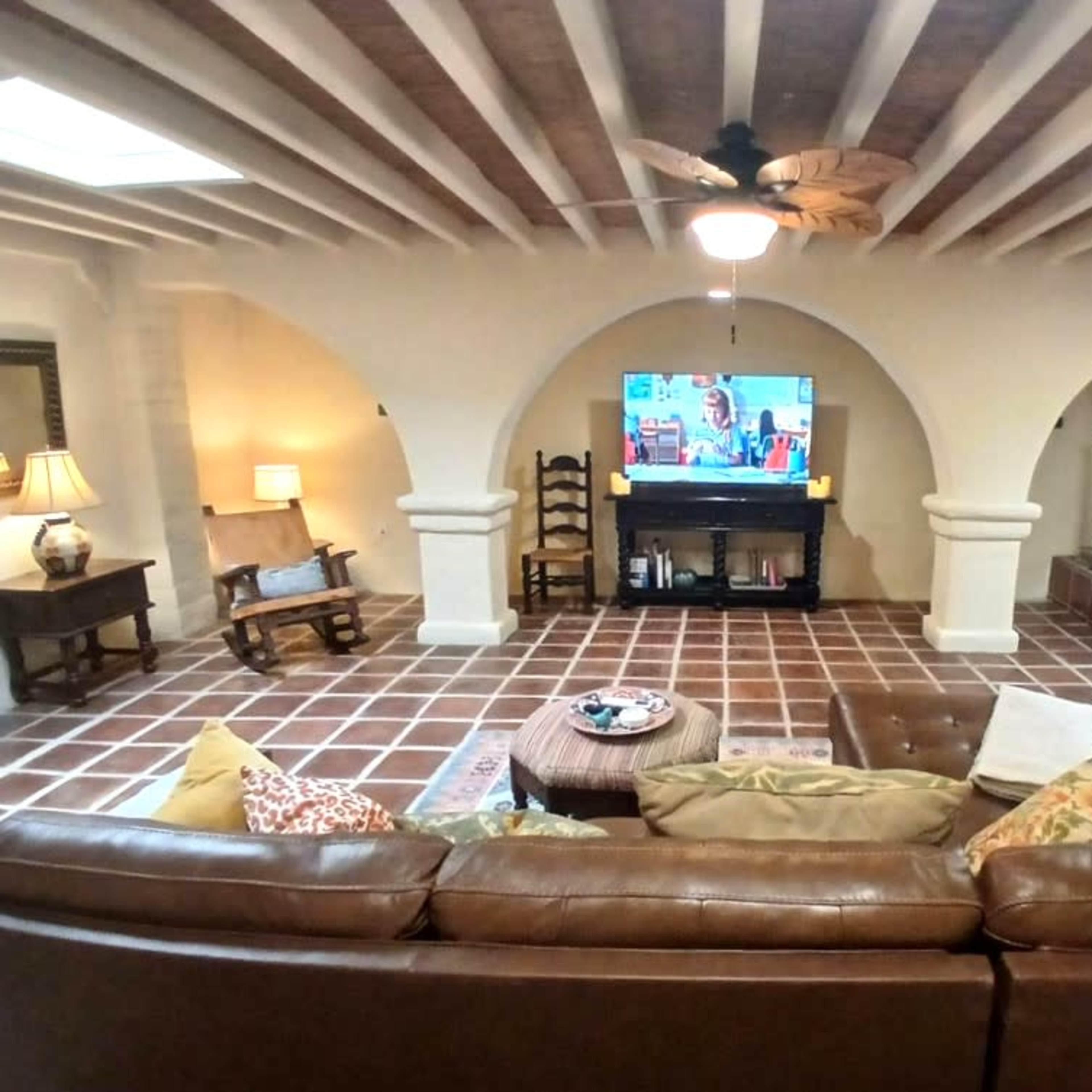 The living room features a leather sofa facing a wall-mounted television, with ceramic tile flooring and arched doorways leading to other spaces.