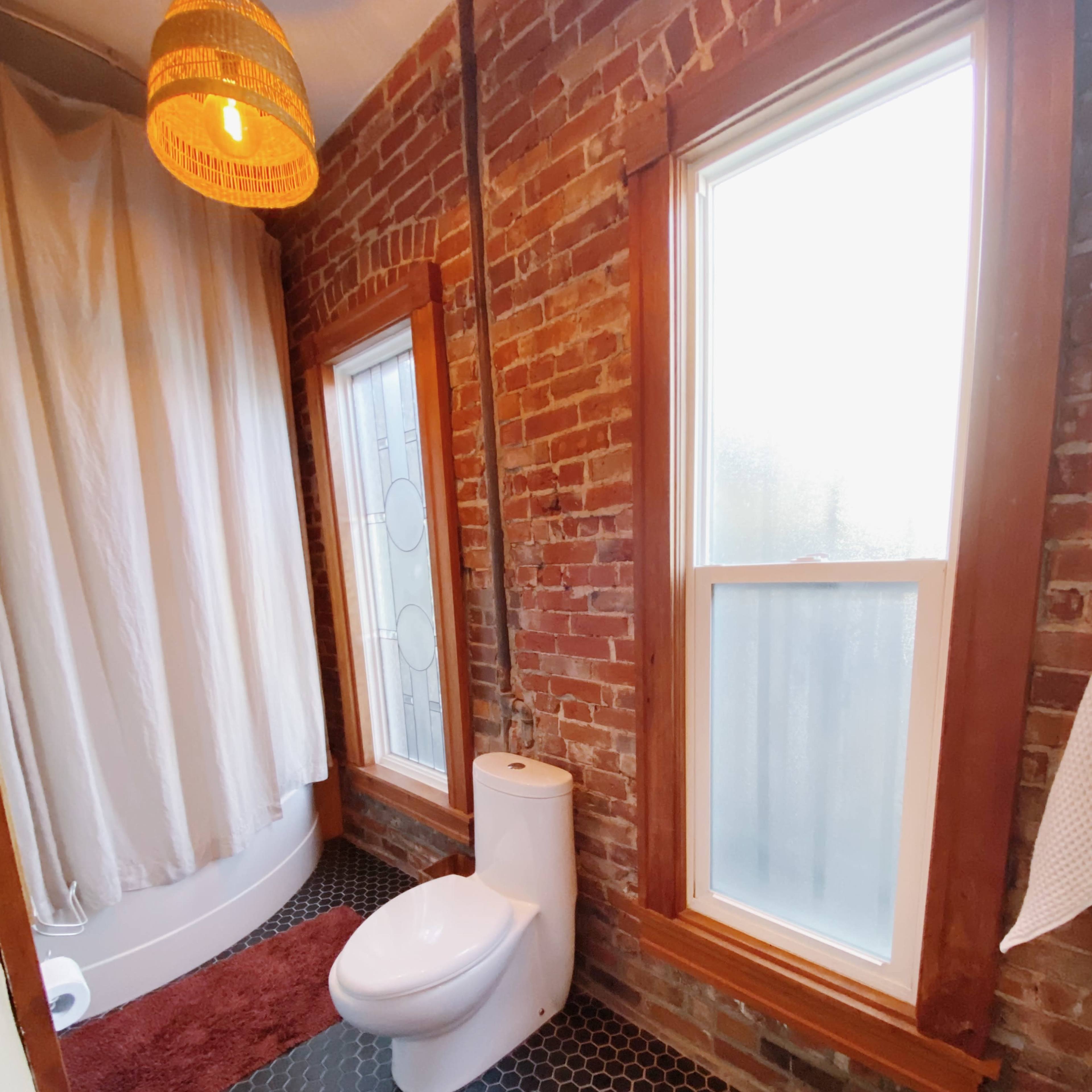 A bathroom with exposed brick walls, a shower behind a curtain, a window, and a toilet on a patterned floor.