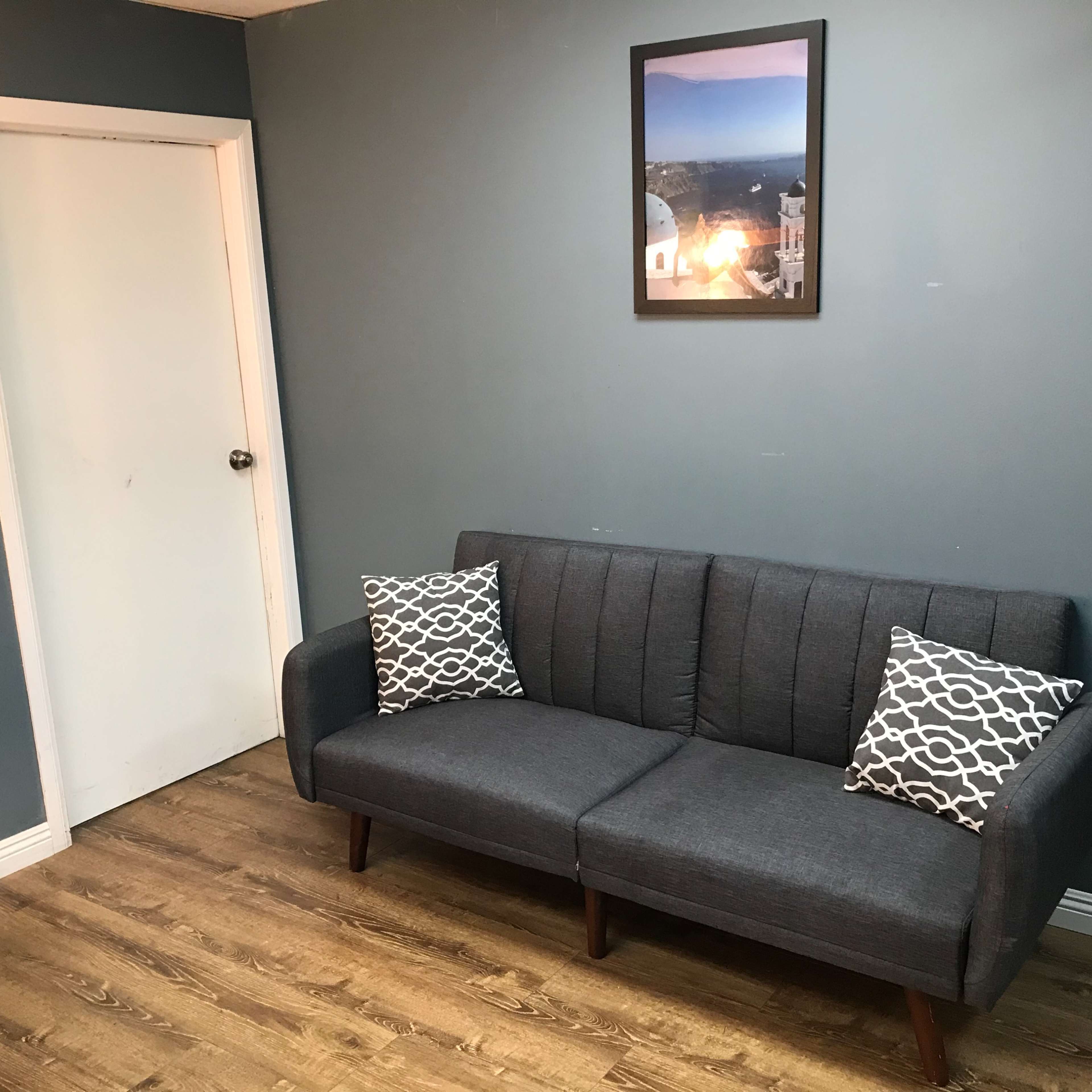 A gray couch with patterned pillows against a blue wall, next to a closed white door and a framed picture hanging above the couch.