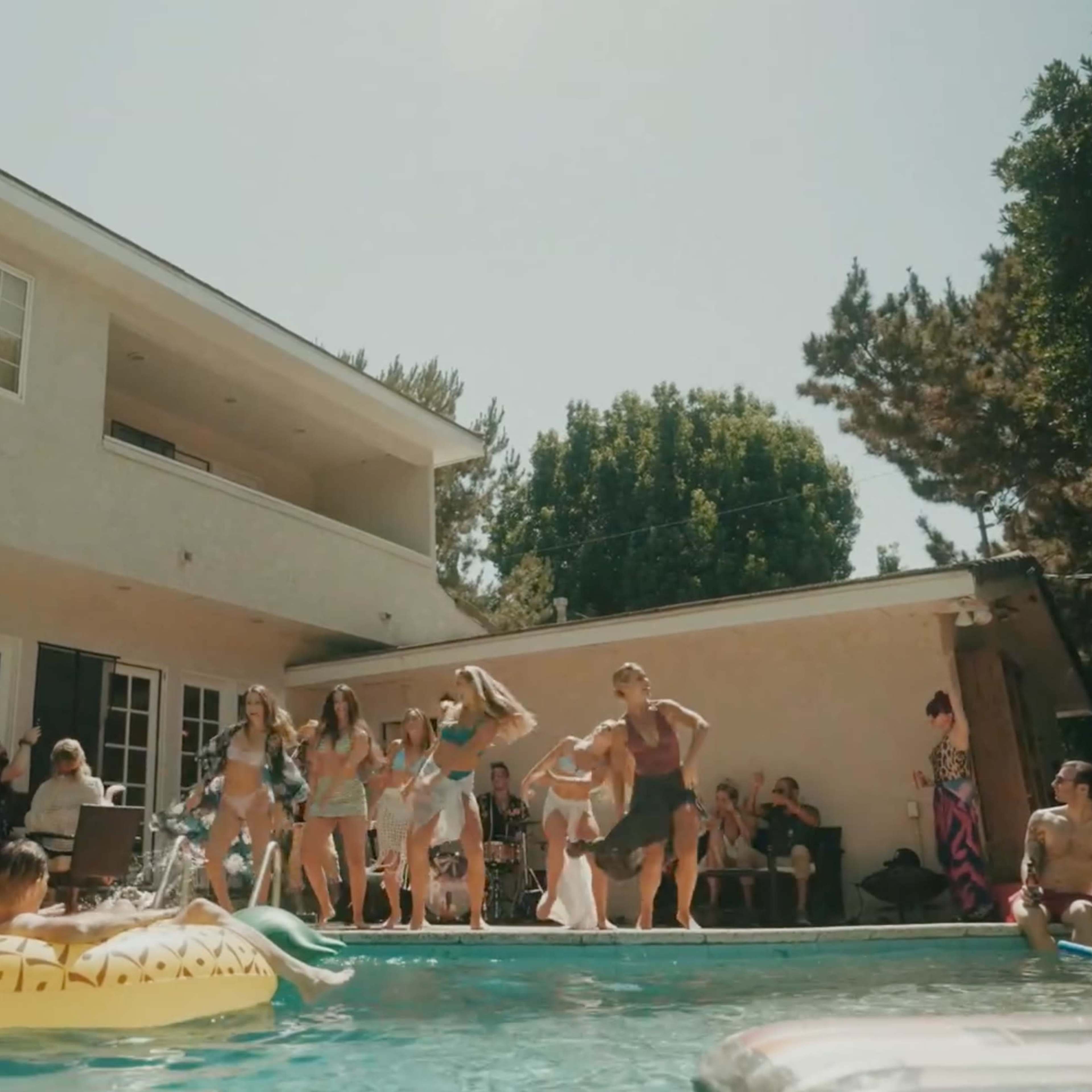 A group of people enjoys a lively gathering by a pool at a house, with some swimming and others socializing on the patio.