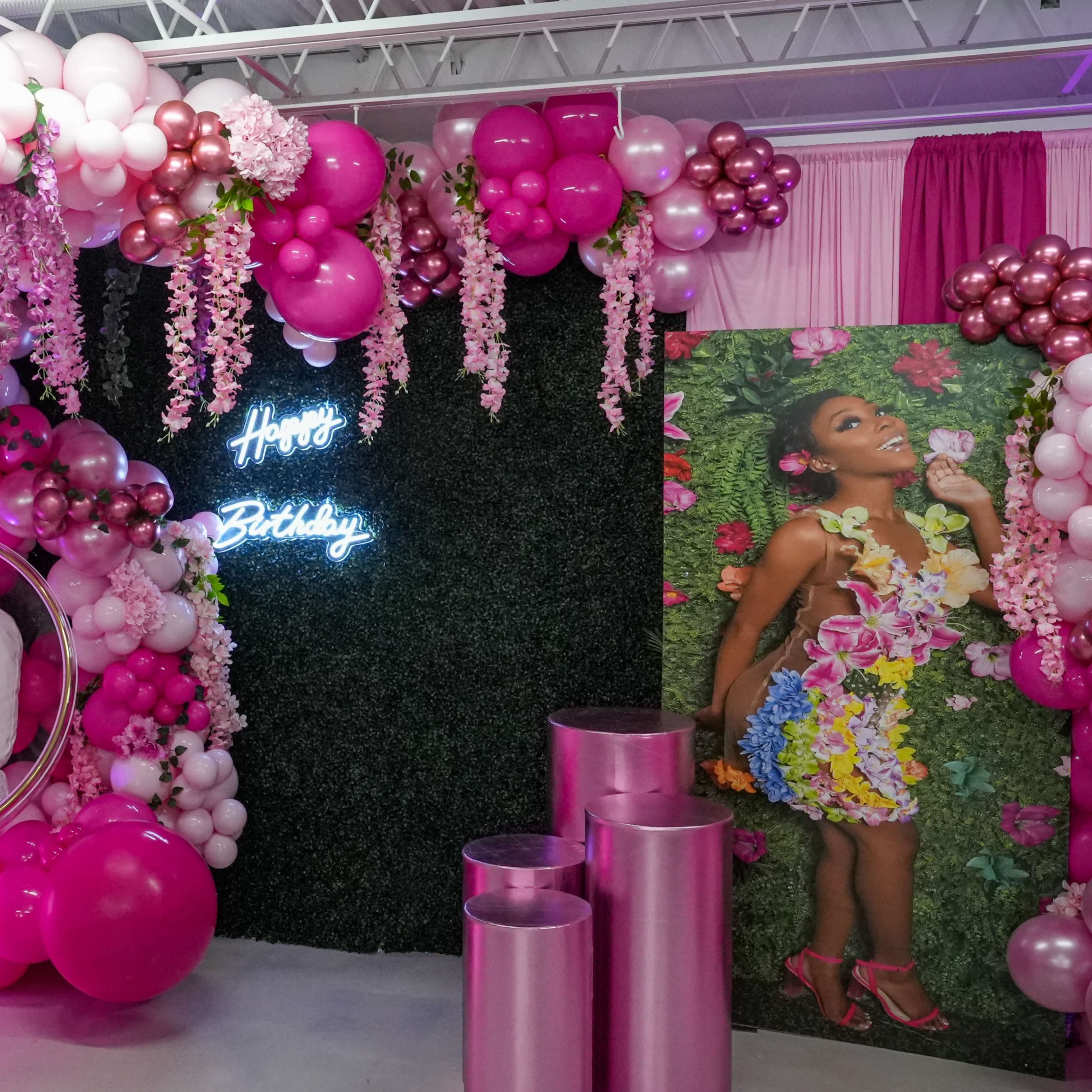 The scene features a colorful birthday decoration with a balloon arch, neon sign, and a floral-themed backdrop.