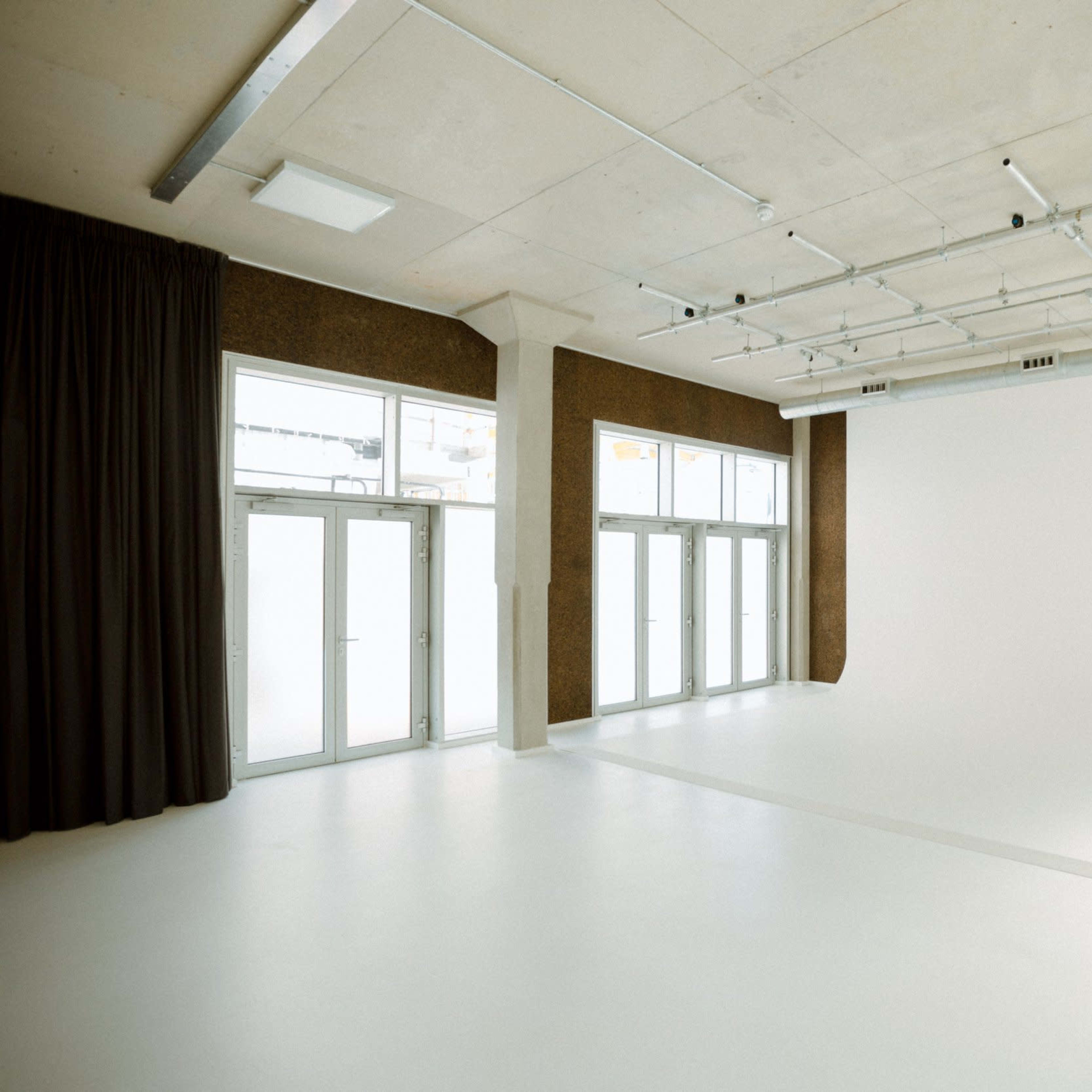 A spacious studio with large windows, white walls, and a blank backdrop, featuring a polished floor and exposed ceiling infrastructure.