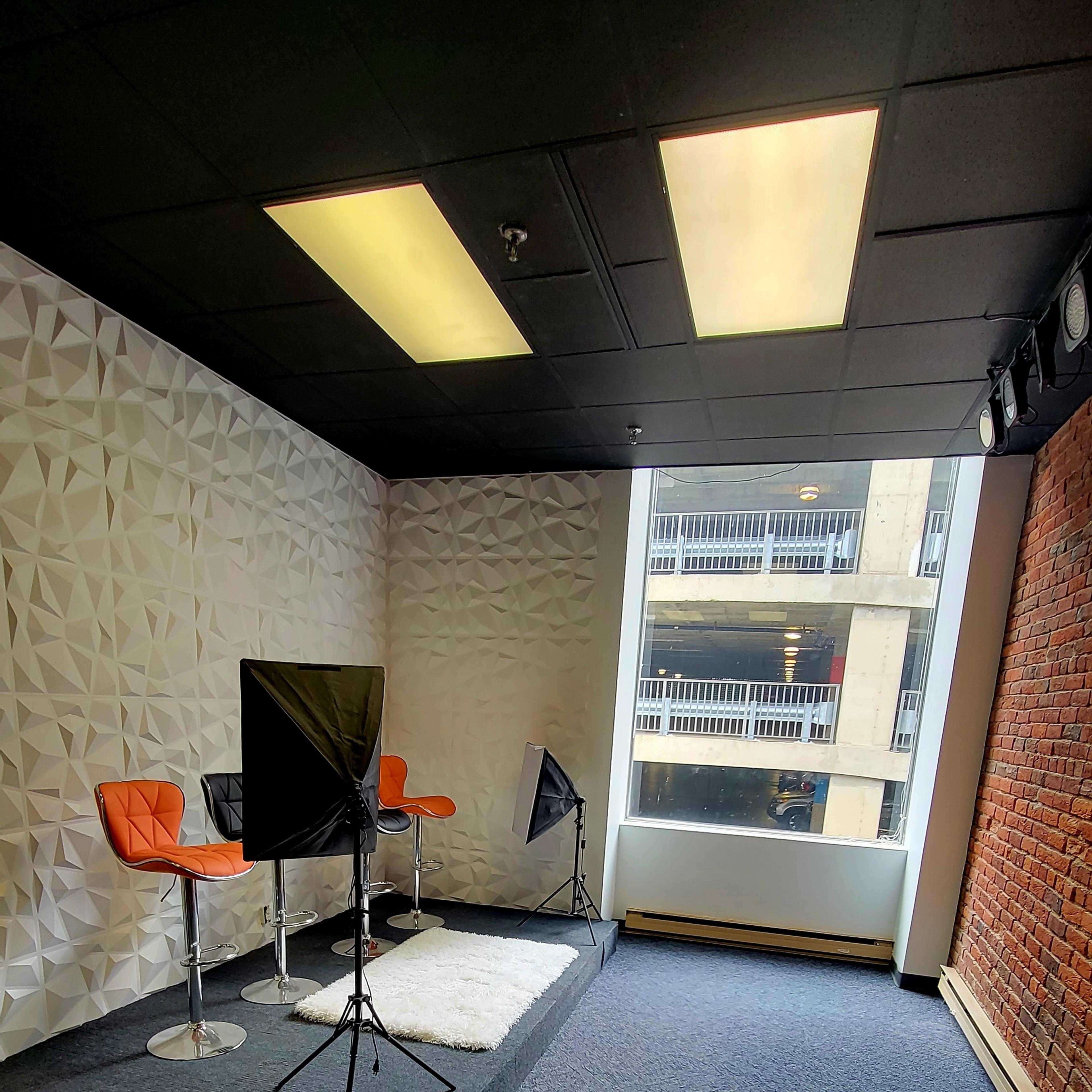 A modern studio space featuring two orange chairs, a black backdrop on stands, and a large window with a view of a building exterior.
