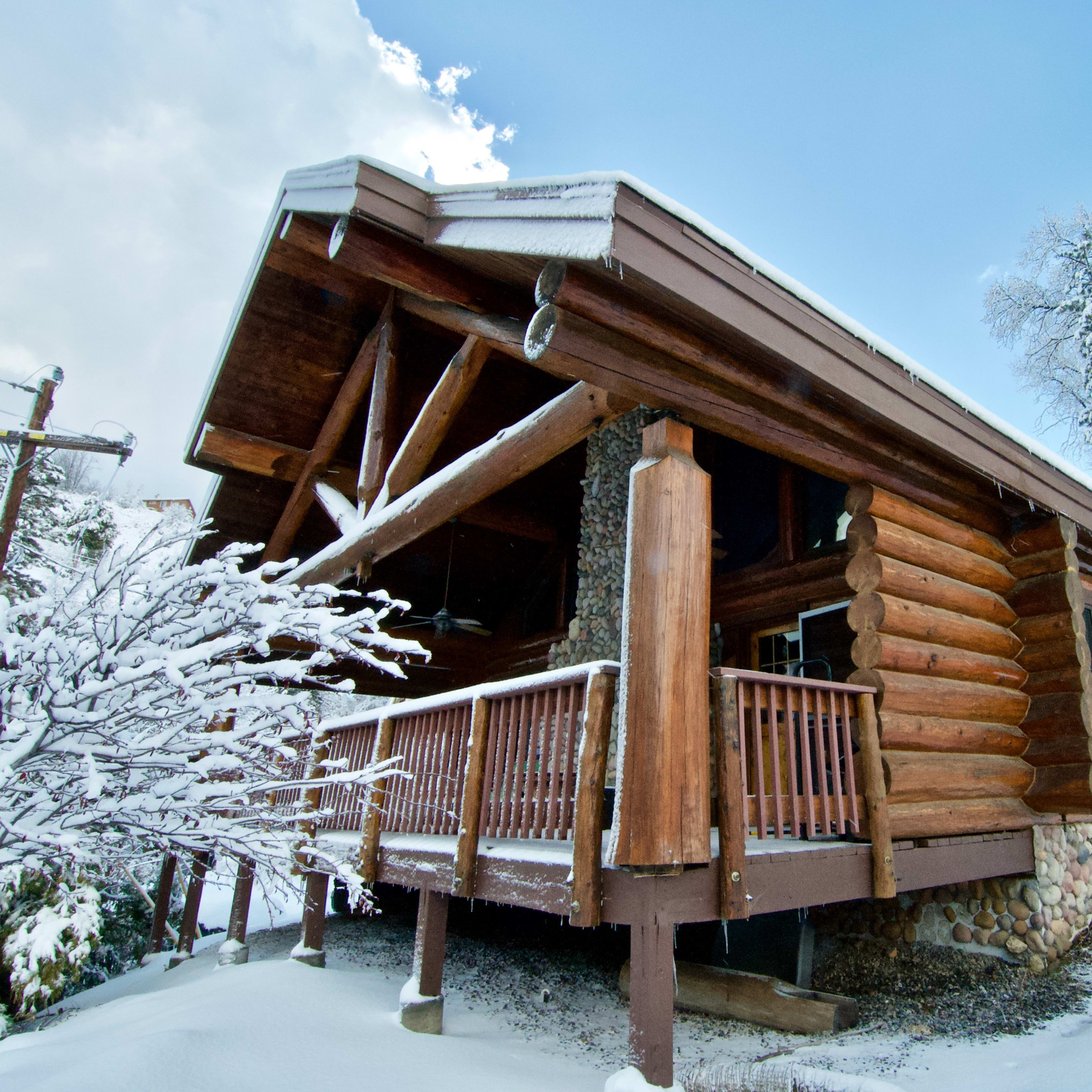 A log cabin with a stone foundation is surrounded by snow-covered trees under a partly cloudy sky.