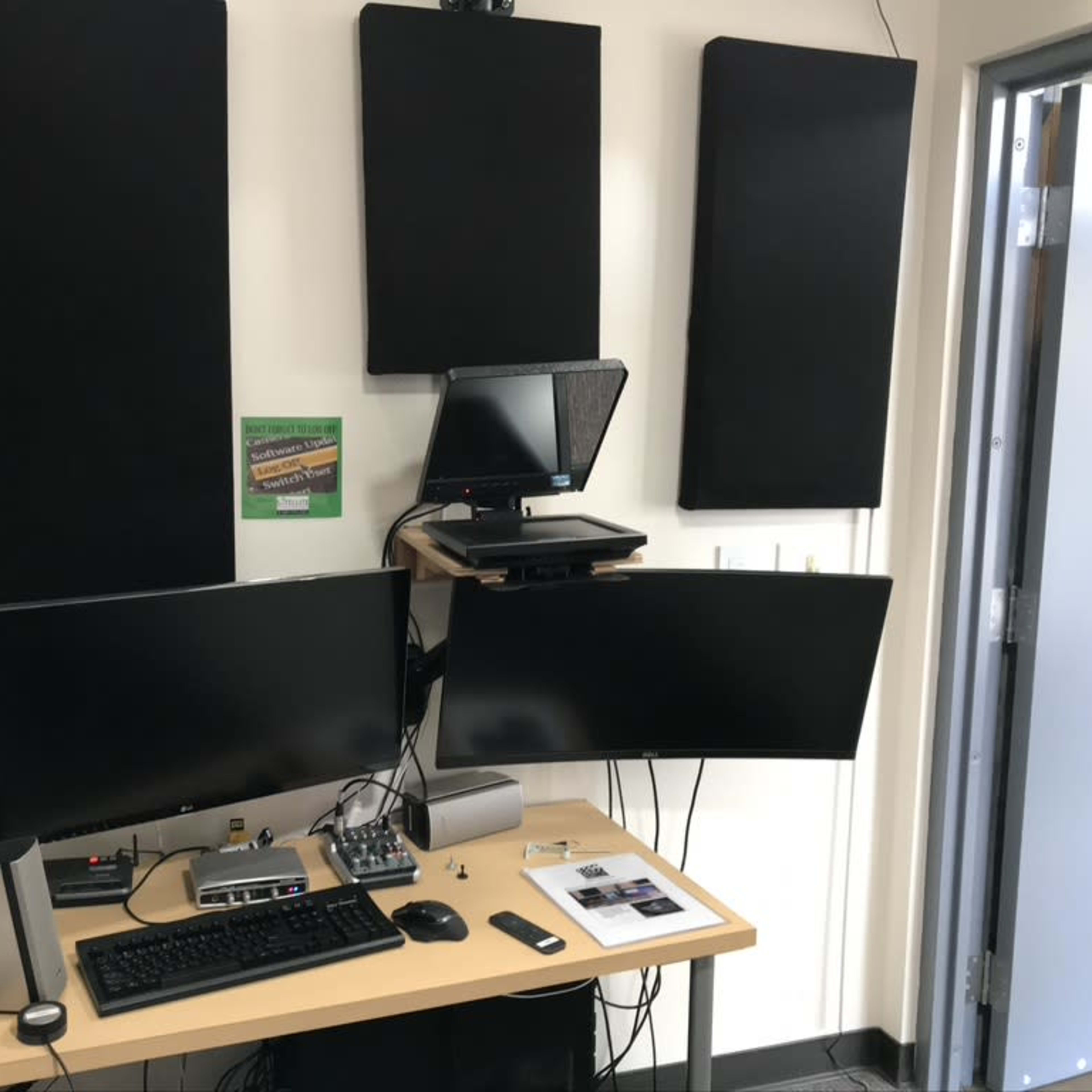 A small workspace with two computer monitors, audio equipment, and soundproofing panels on the wall.