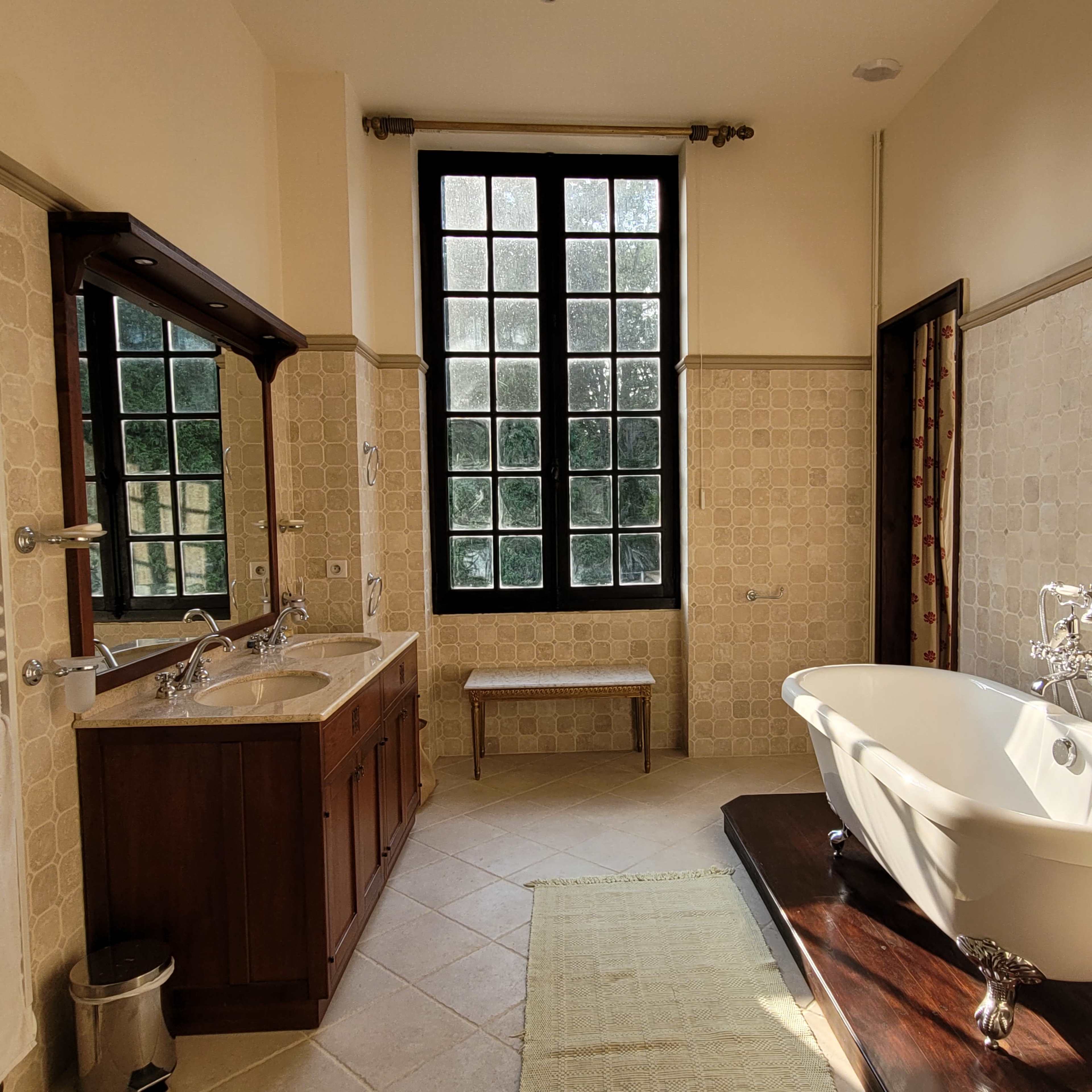 A spacious bathroom featuring a freestanding bathtub, double sink vanity, a large window, and tiled walls.