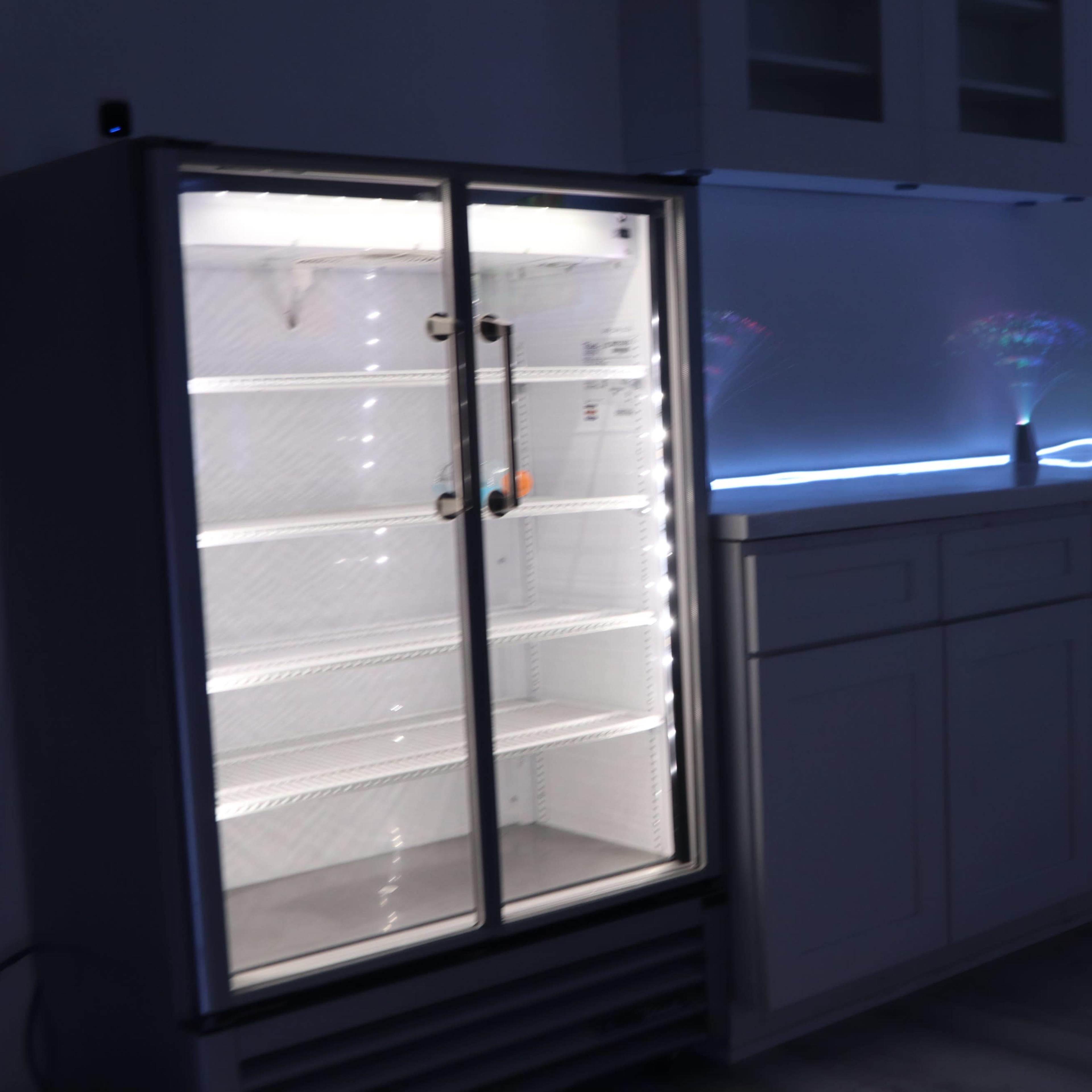 An empty refrigerator with glass doors next to a white cabinet in a dimly lit room.