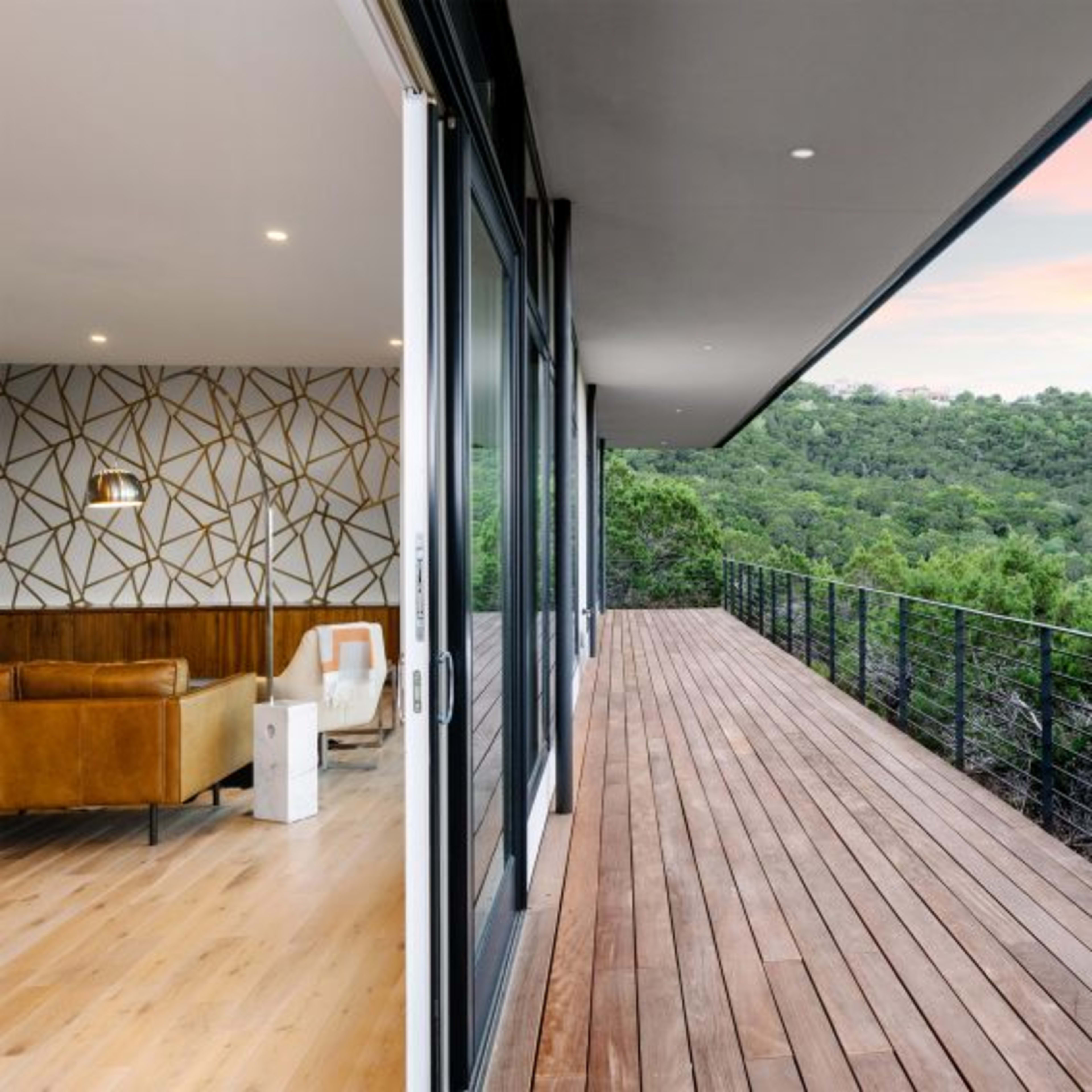 A wooden deck extends from a modern interior, offering views of a lush green hillside and a colorful sky.