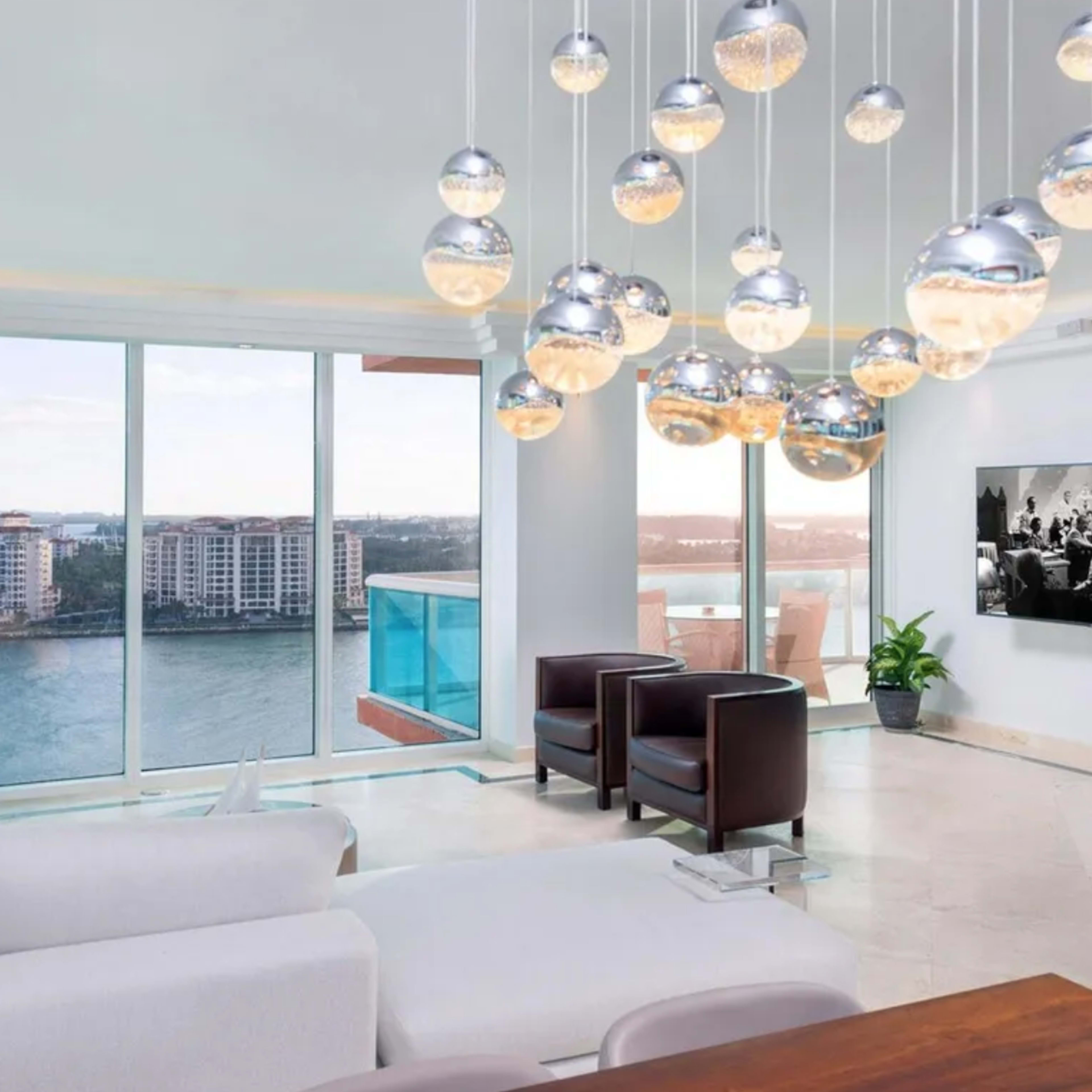 A modern living room with a large window displaying a waterfront view, two brown armchairs, a white sofa, a wooden dining table, and pendant lighting overhead.