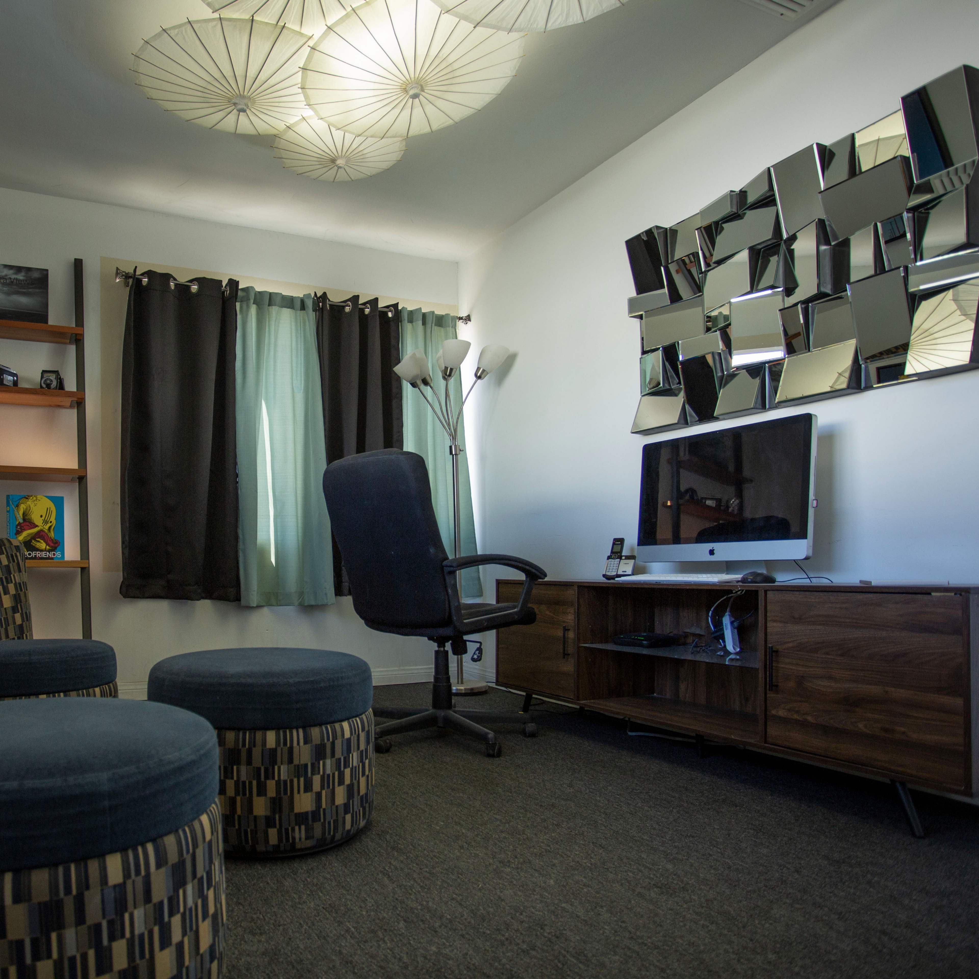 A small living space features two patterned armchairs, a round ottoman, a desk chair, a television on a wooden stand, and decorative lighting above.