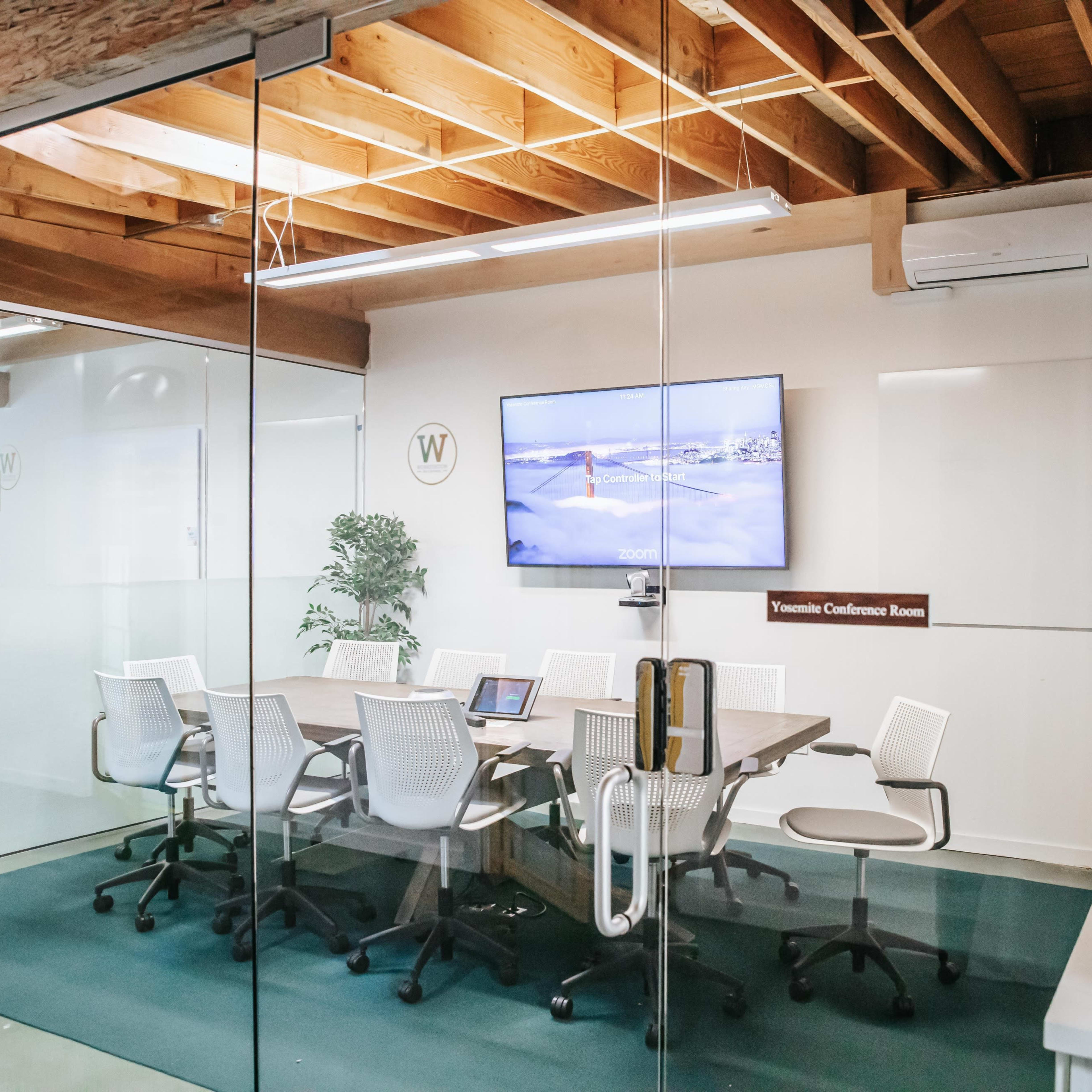 A modern conference room with a glass wall, a large screen on the wall, a table surrounded by six white chairs, and a small counter with a sink.